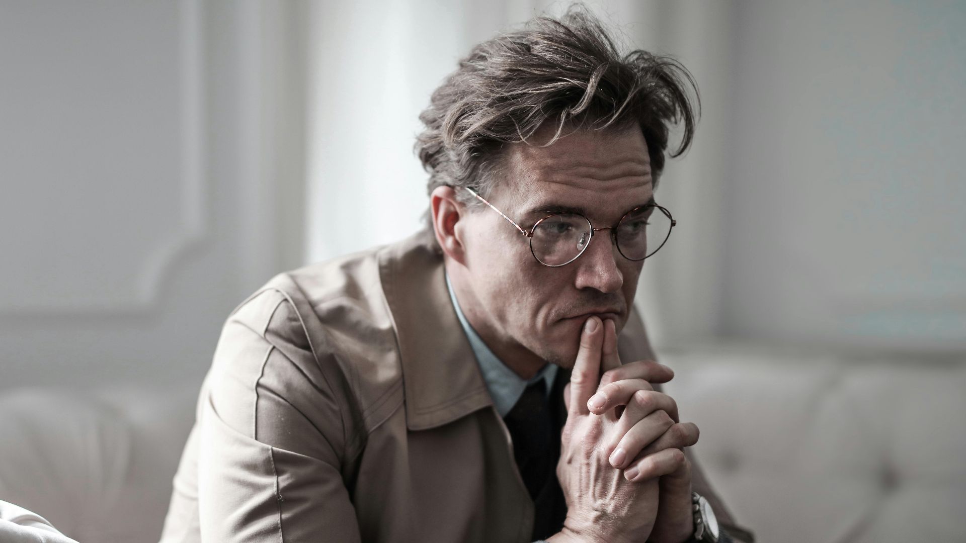 A thoughtful man wearing glasses sits on a sofa in a classic interior, lost in deep contemplation.