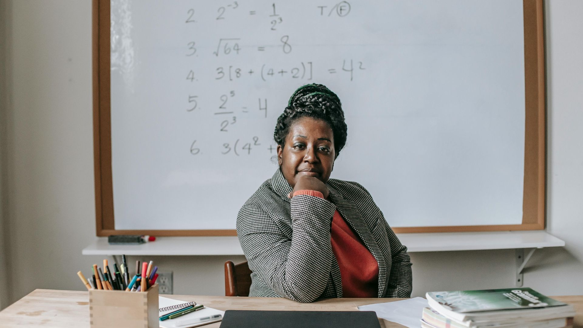 African American female teacher in formal clothes sitting at table with books and laptop and looking at camera