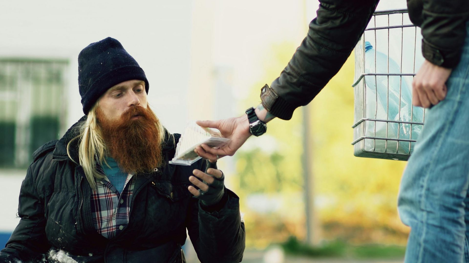 A bearded homeless man receives food from a passerby outdoors.