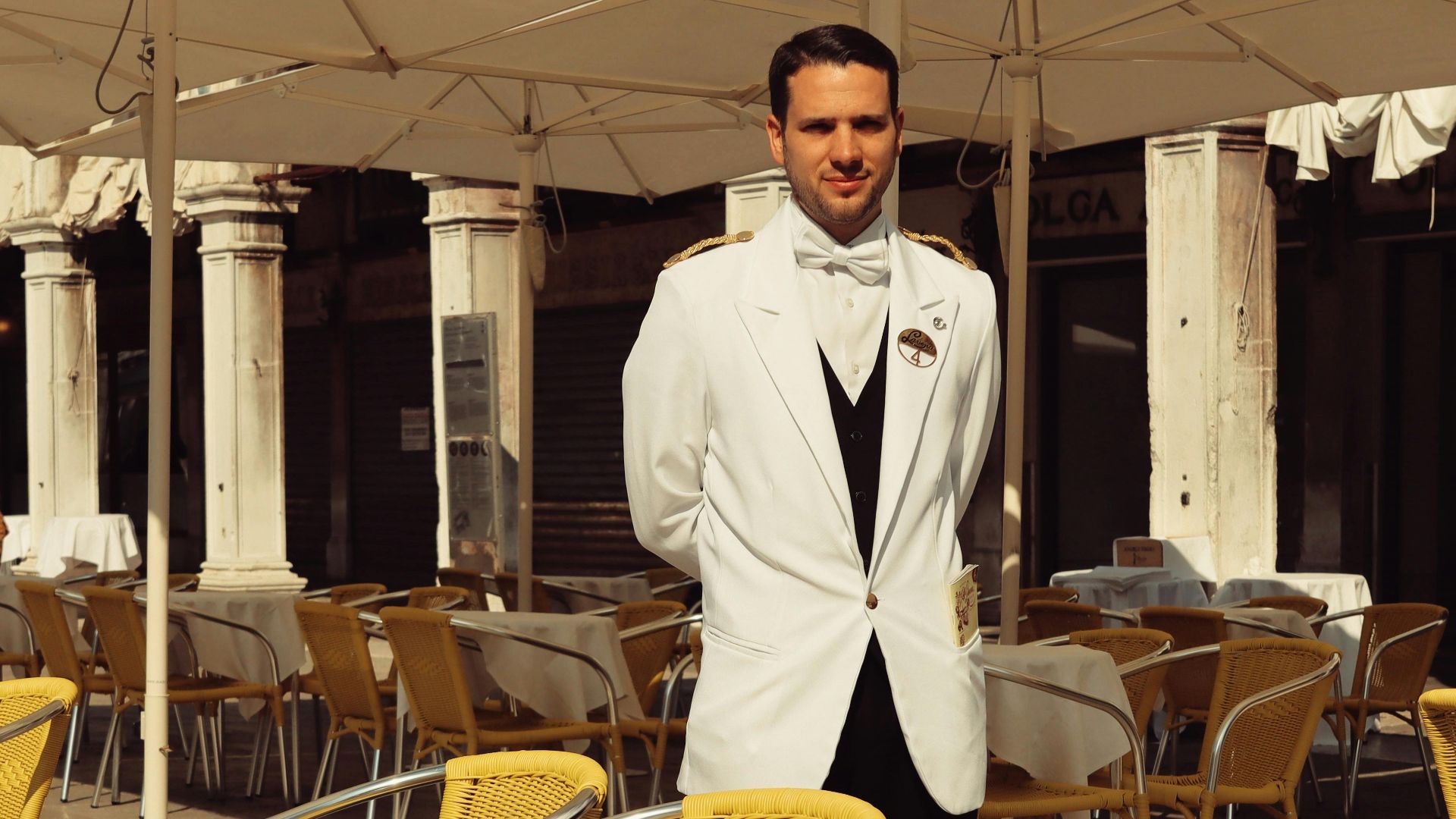 A waiter in a white suit stands amidst sunlit tables in a Venice café.