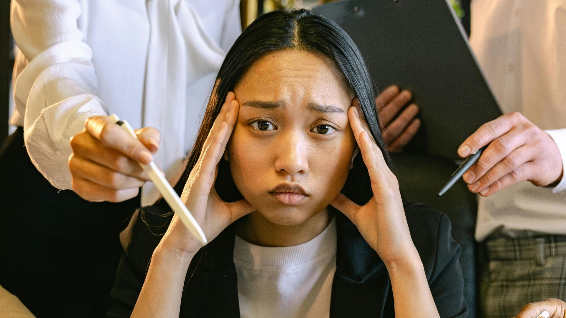 A stressed young woman overwhelmed with work at the office, surrounded by colleagues and devices.