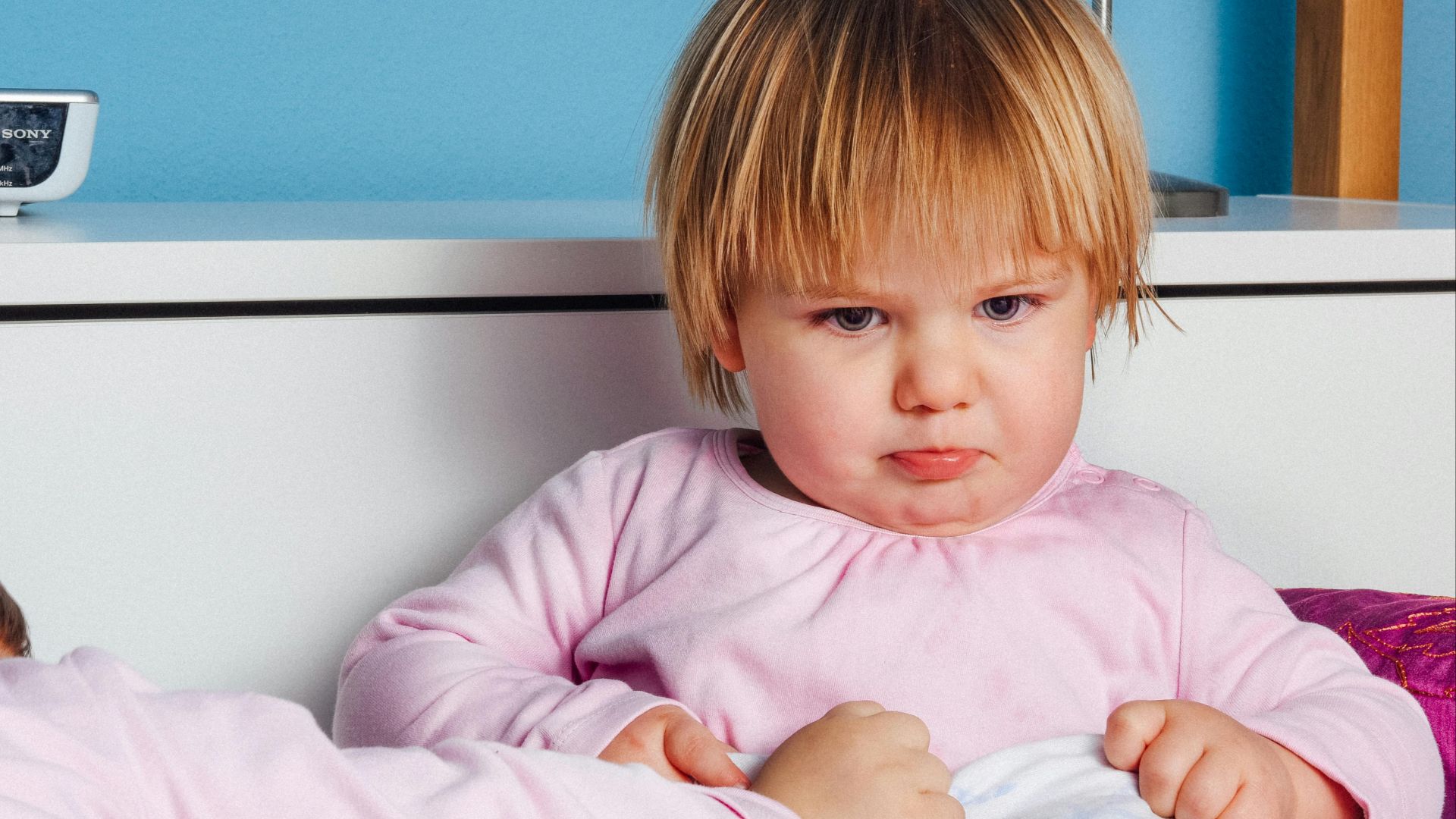 Cute toddler with a pout in a pink outfit indoors, showcasing an expressive mood.
