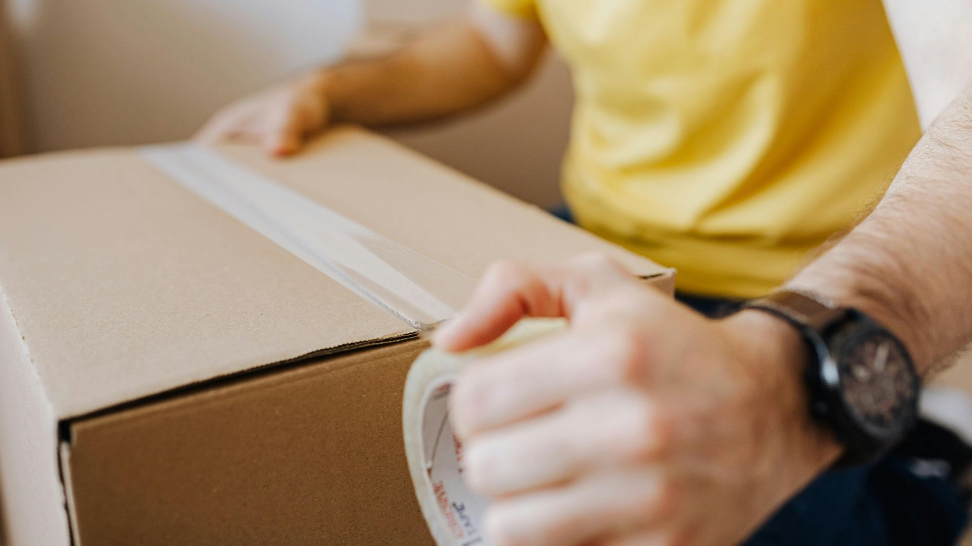 Crop anonymous male in casual wear using cling tape to seal cardboard container with belongings during relocation