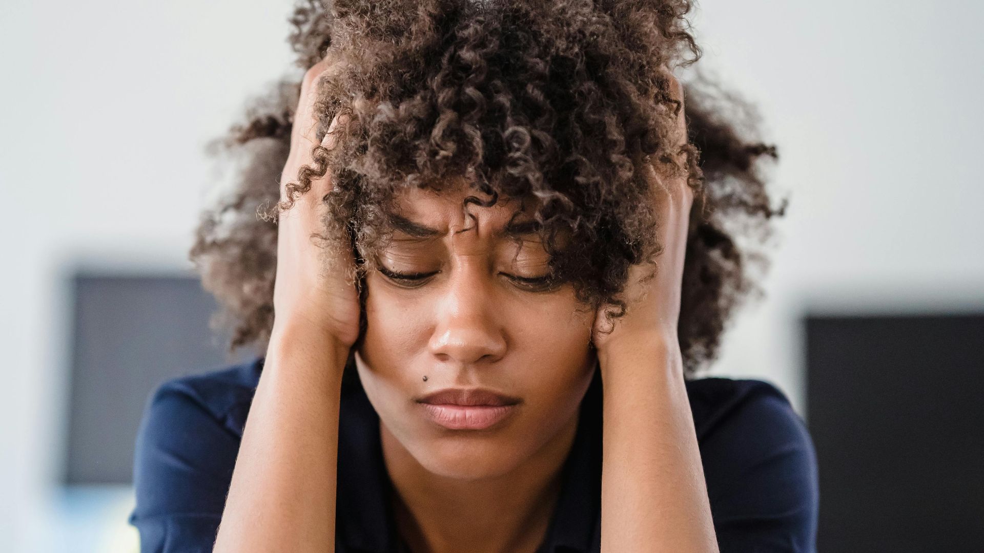 Woman in office setting expresses stress, seated at desk with hands in hair. Perfect for workplace stress concept.