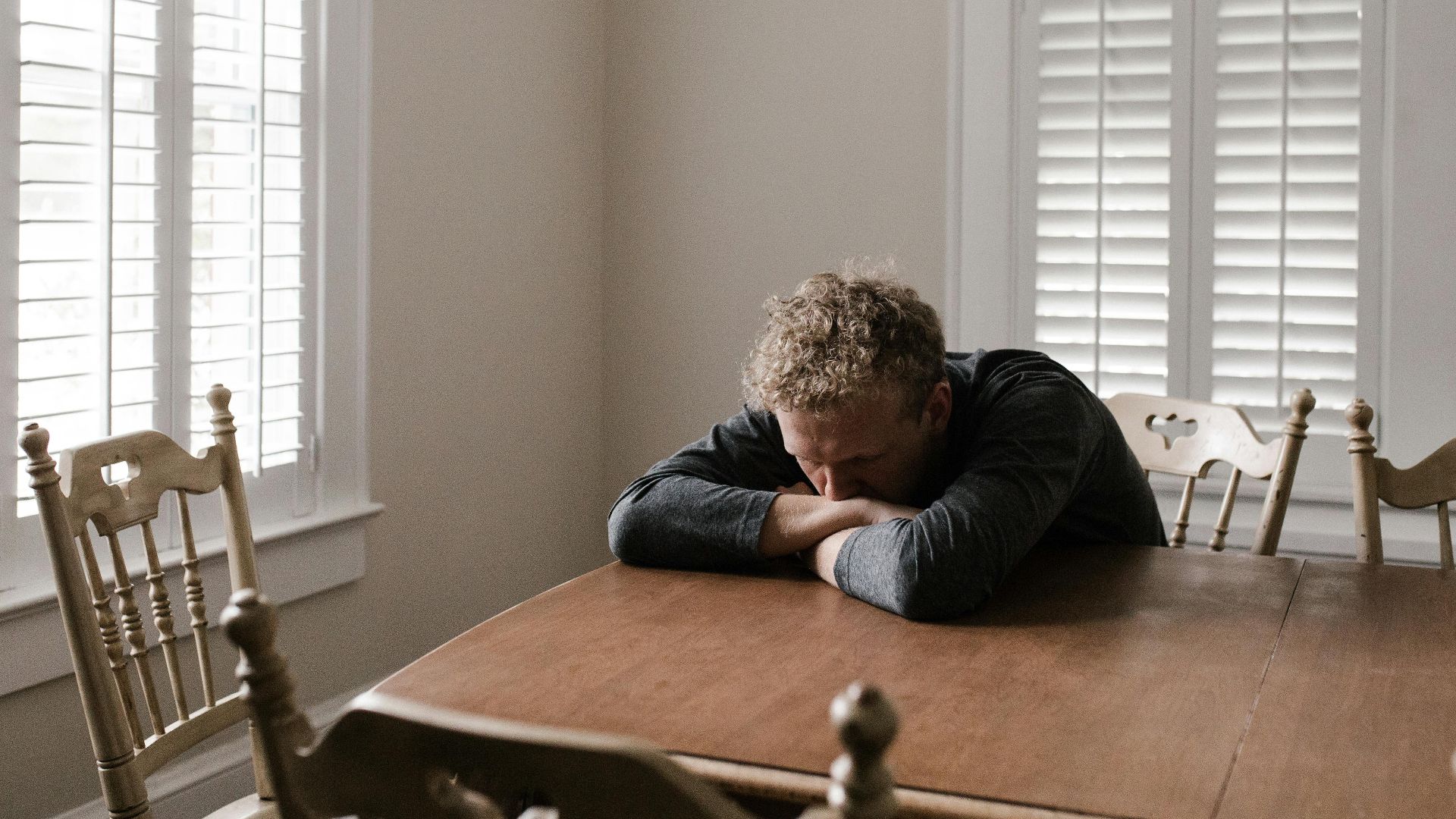 An adult man with emotions of sadness and anxiety sitting alone at a wooden table near windows.