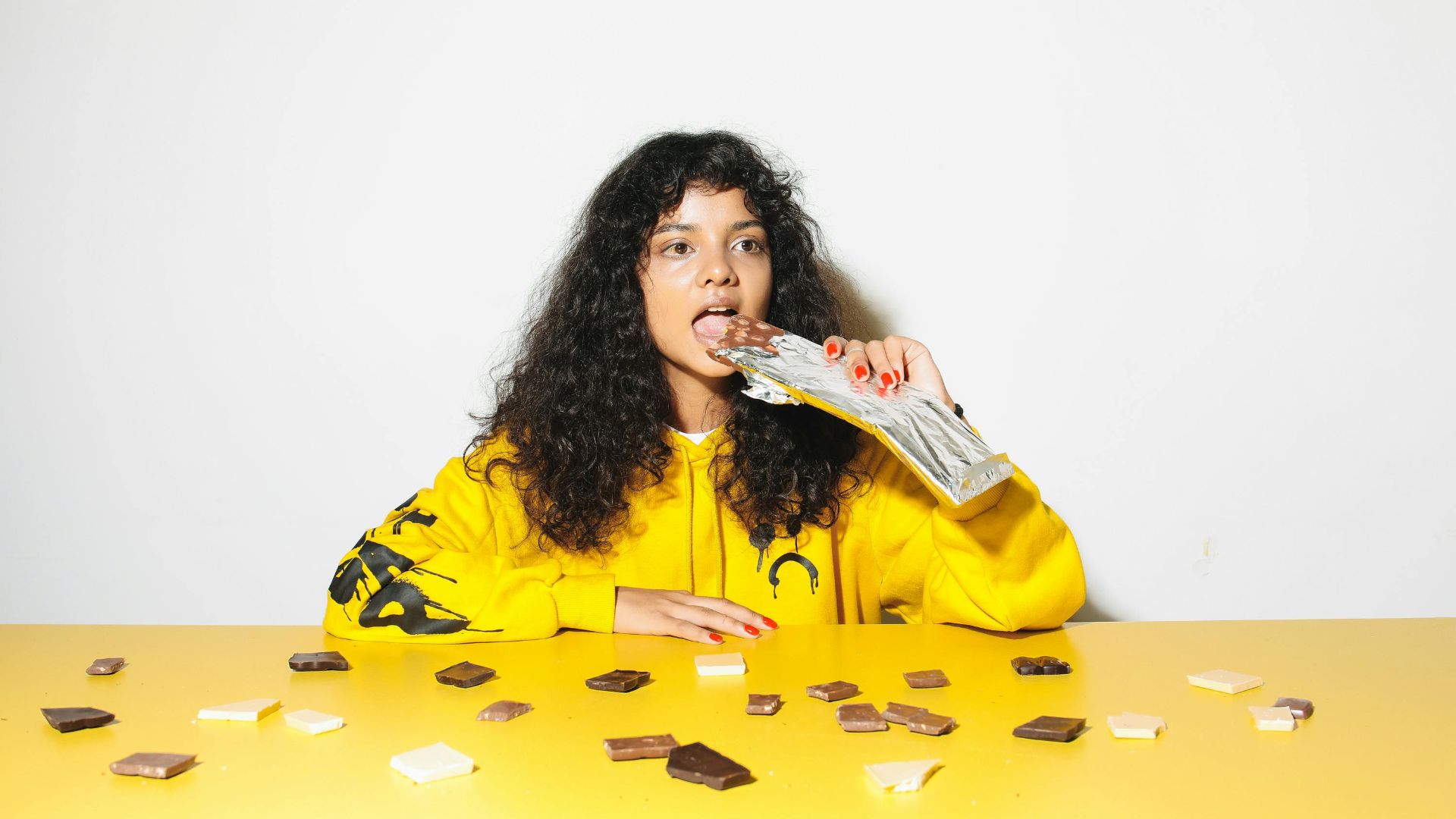 A young woman in a yellow hoodie enjoying chocolate pieces at a table. Bright scene.