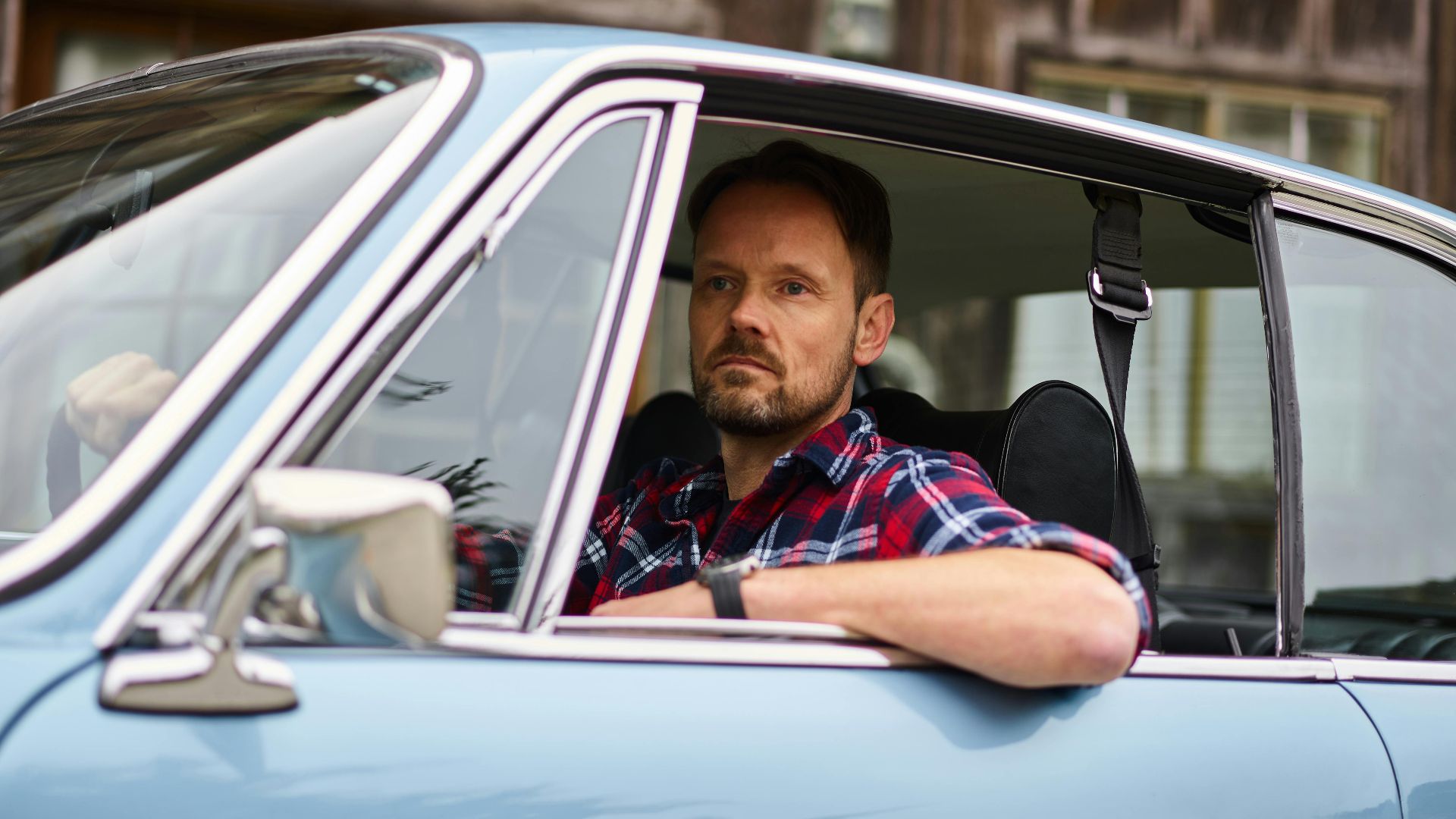 Adult thoughtful bearded driver in wristwatch and casual clothes driving blue automobile while passing by old building and looking away in daylight