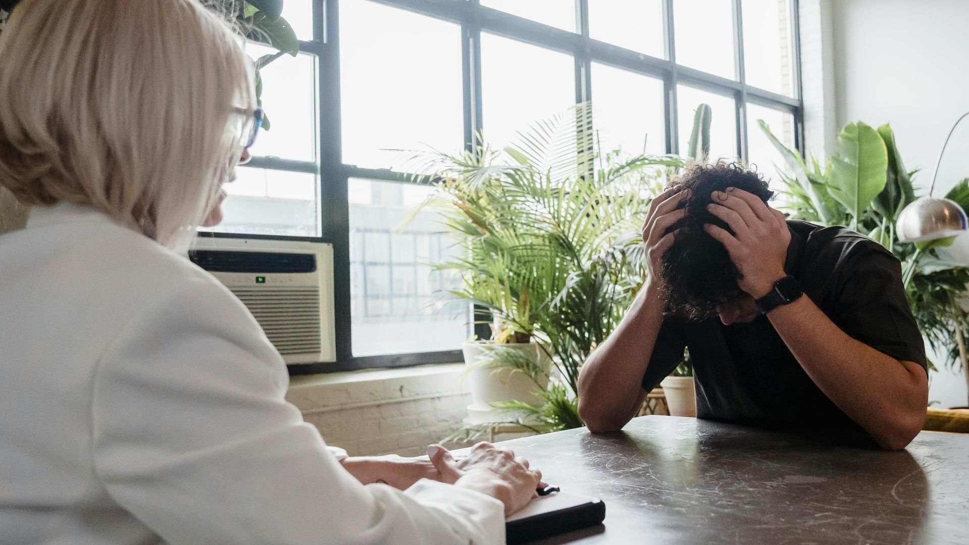 A counselor listening to a stressed client in a plant-filled, modern office.