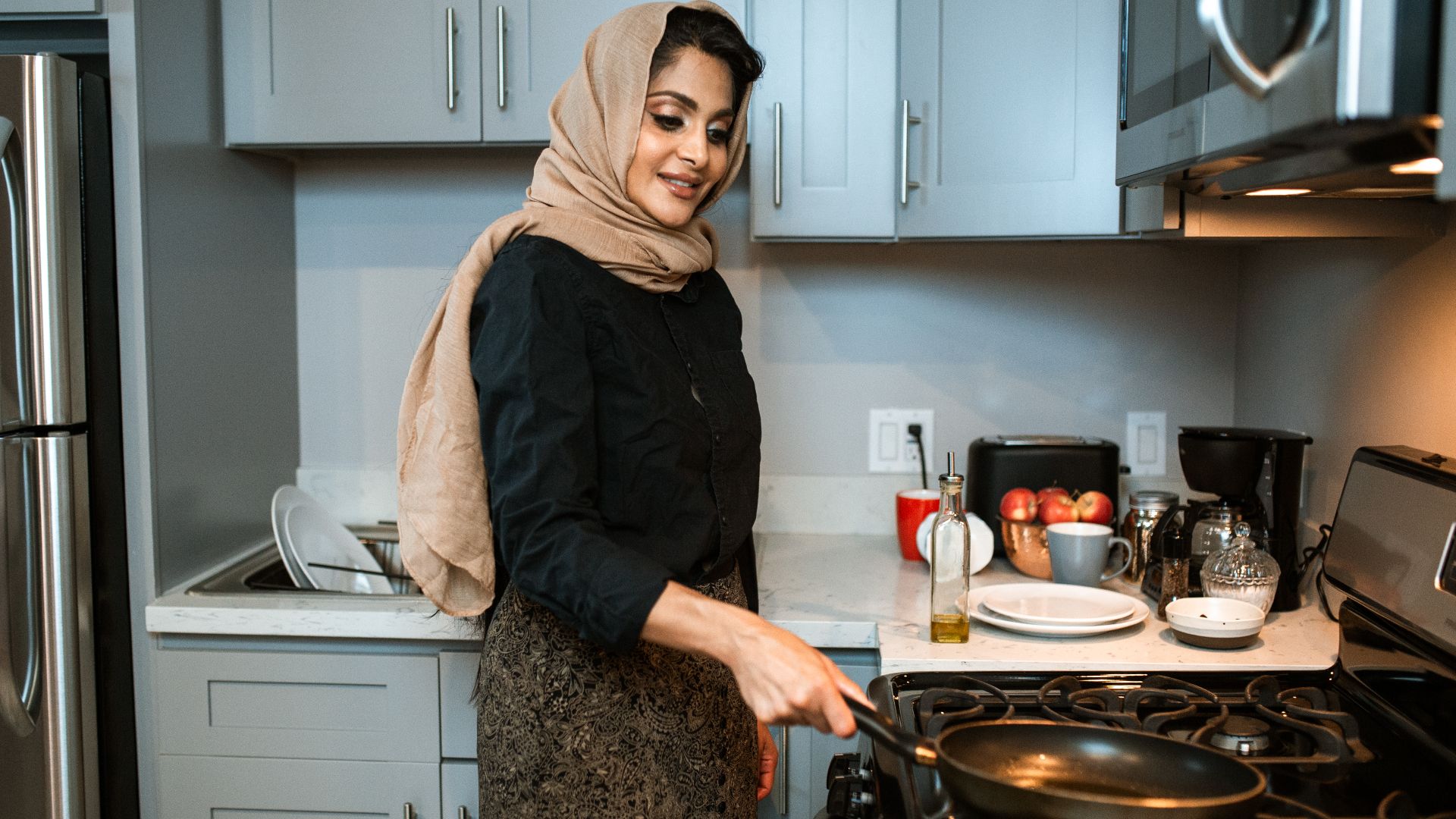 Positive ethnic female wearing headscarf and stylish clothes putting frying pan on gas stove while preparing lunch in contemporary light kitchen