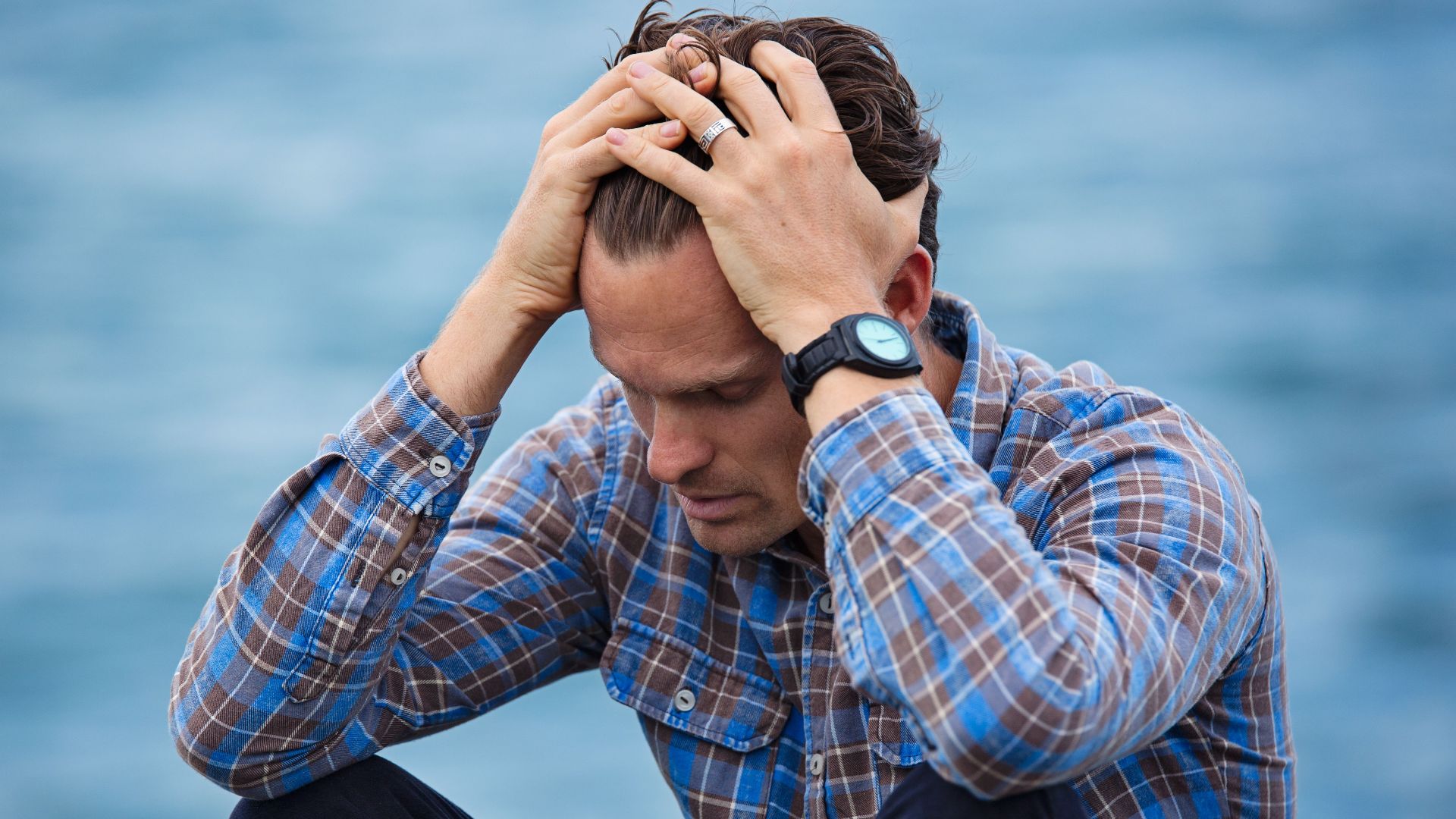 A man in a plaid shirt sits by the water looking distressed, symbolizing stress.