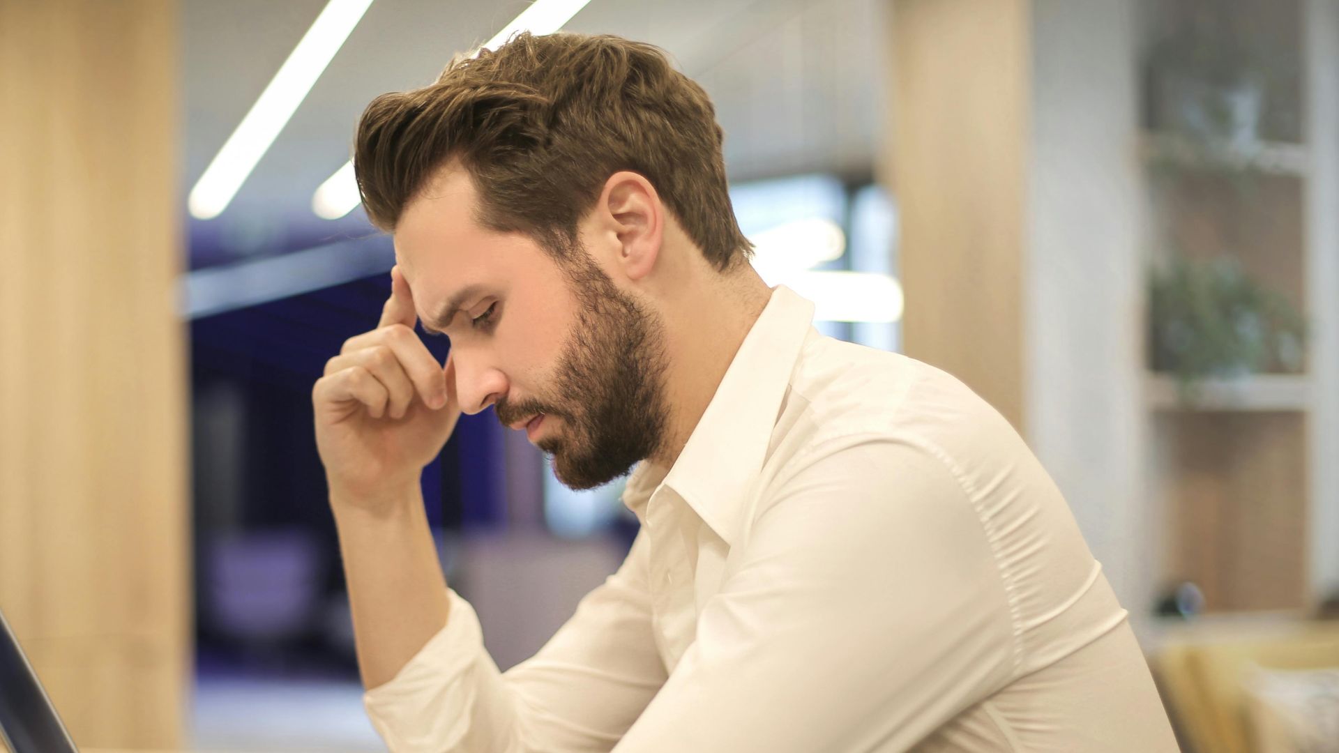A thoughtful man in a shirt works on his laptop at a modern indoor office space.