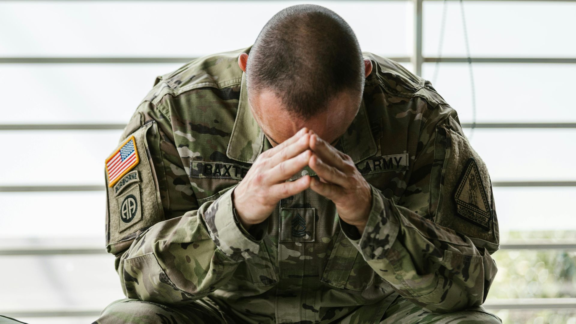 A soldier in camouflage sitting indoors, appearing stressed and overwhelmed.