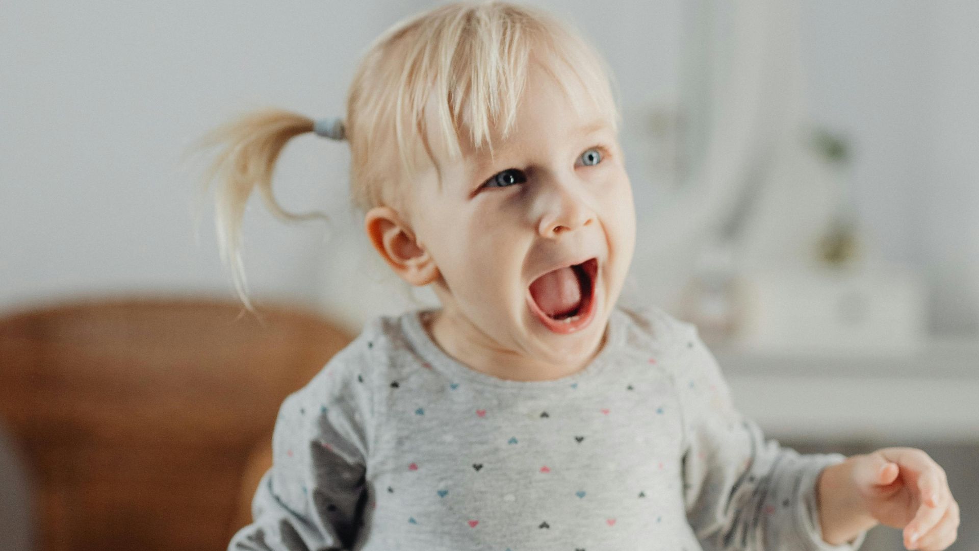 A happy blonde toddler wearing a gray dress, smiling joyfully indoors with a blurred background.