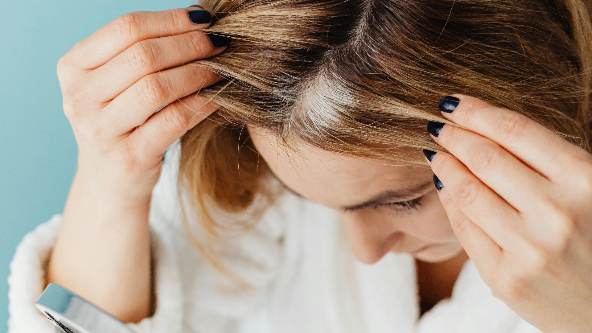 Woman in bathrobe checking grey hair roots in front of a mirror indoors.