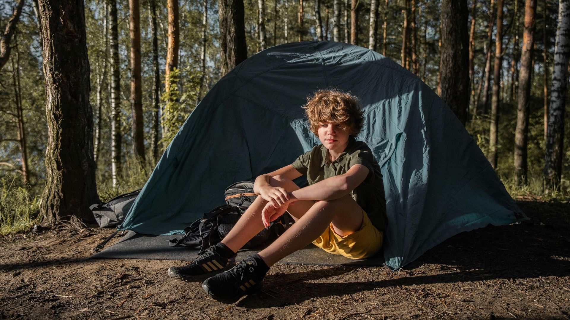 A young boy enjoying a camping trip in a forest, sitting in front of a tent.
