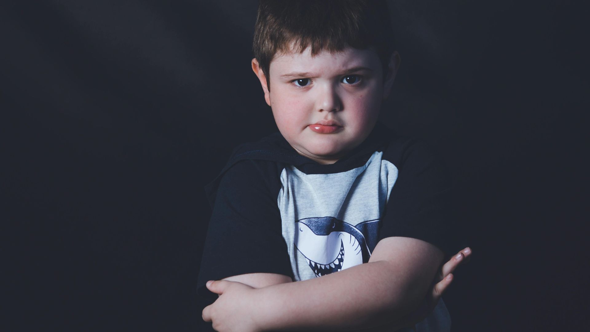 A thoughtful young boy with crossed arms against a dark background, expressing introspection and emotion.