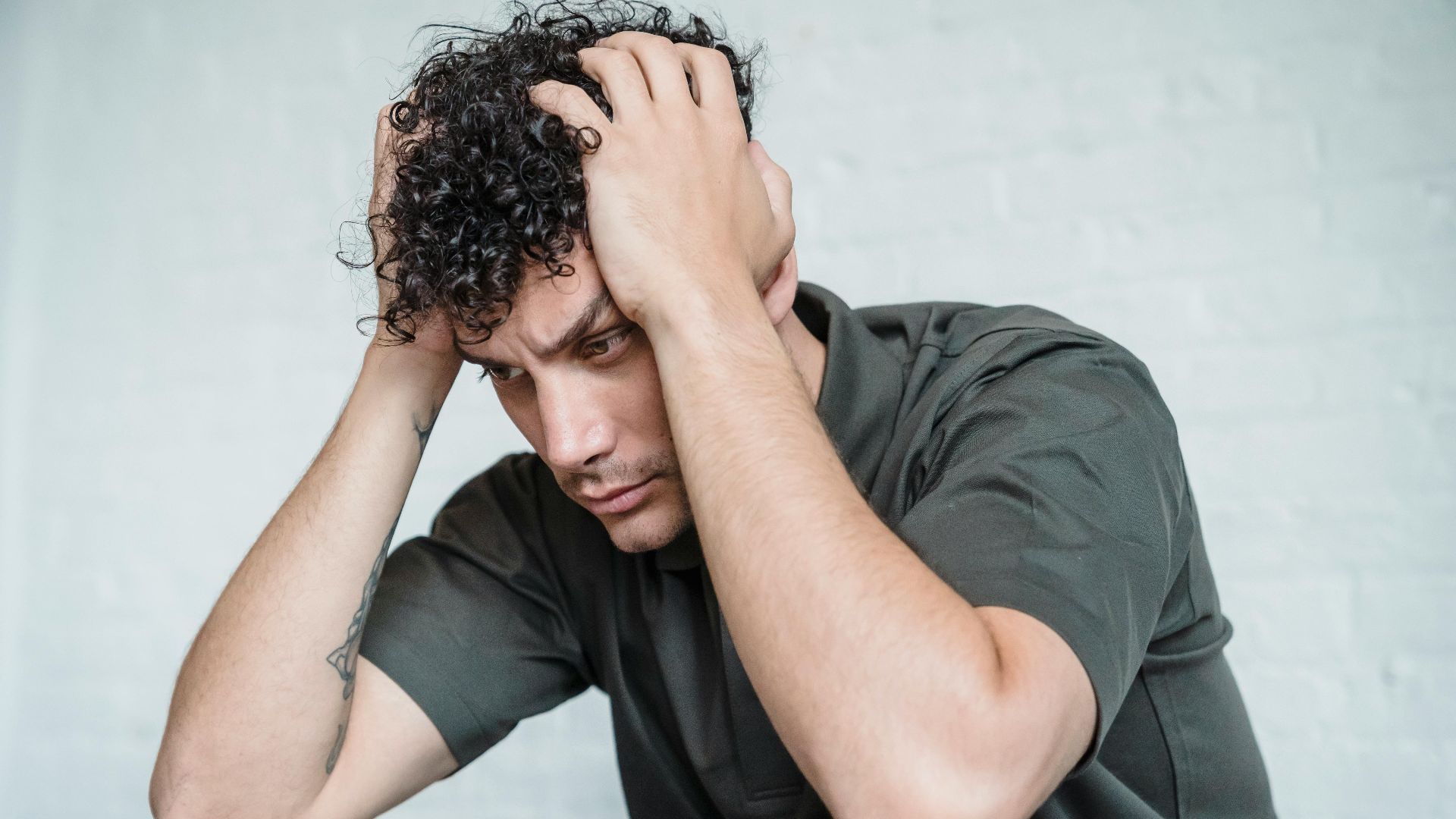 A young man with curly hair looks stressed as he sits with his hands on his head in contemplation.