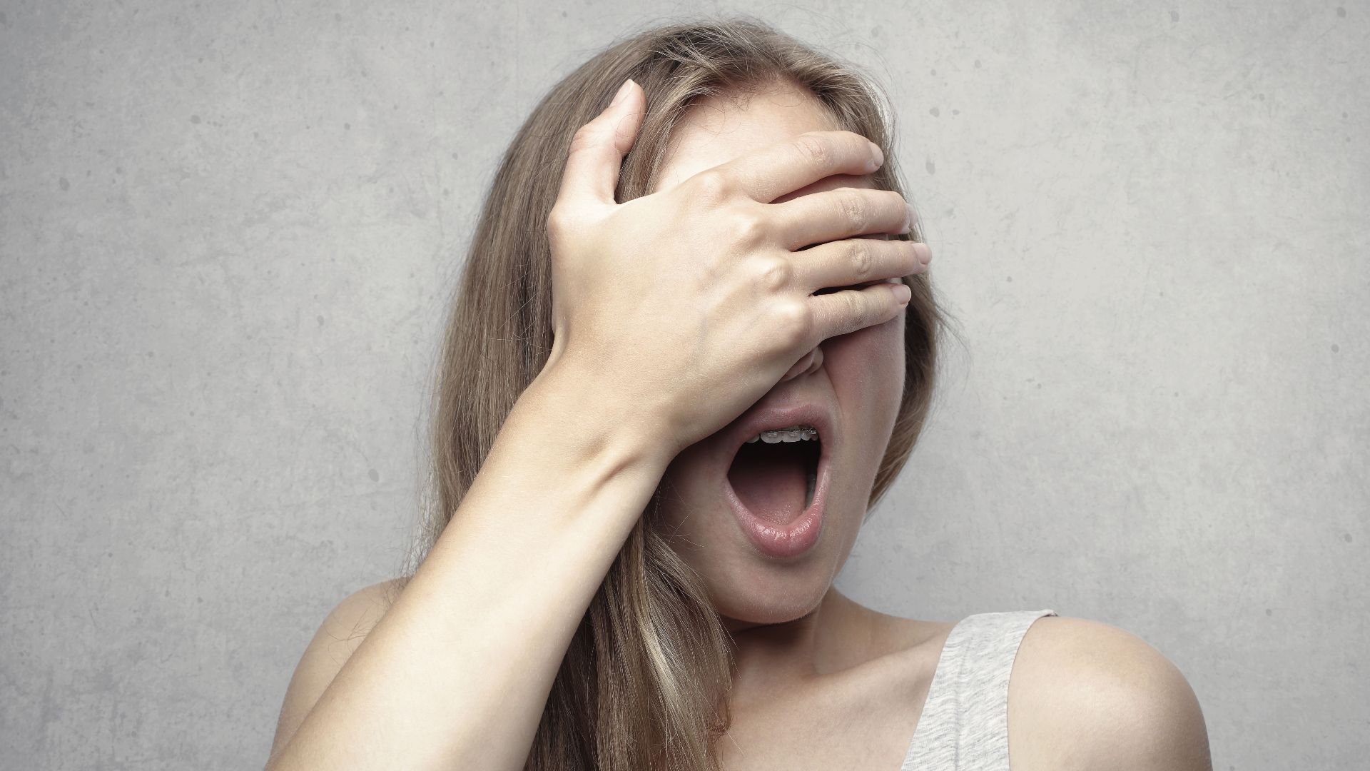 A surprised woman covers her face with one hand, expressing shock and amazement against a gray background.