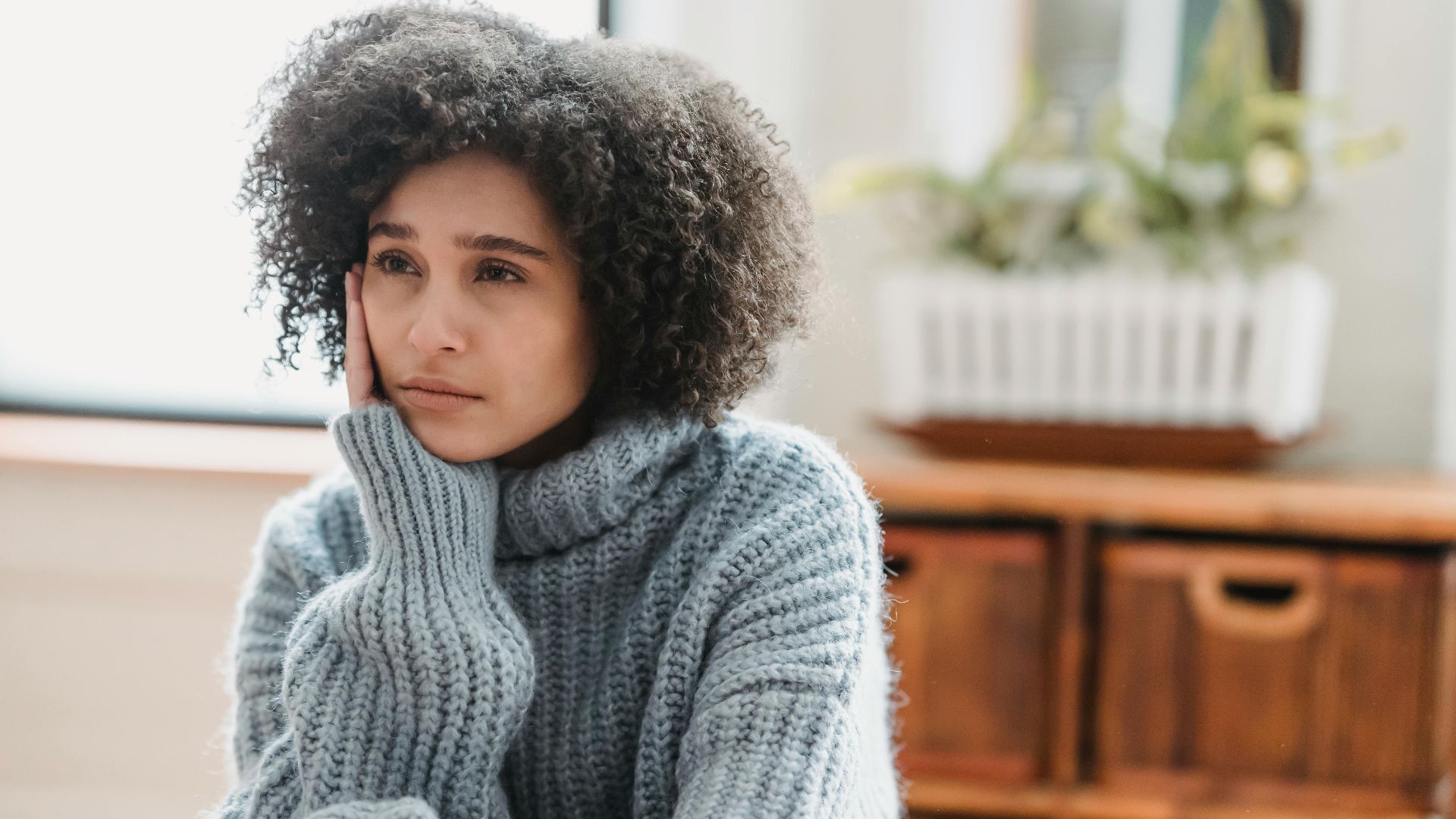 Discontent African American female leaning on hand and looking away while sitting in light room near green plant on blurred background