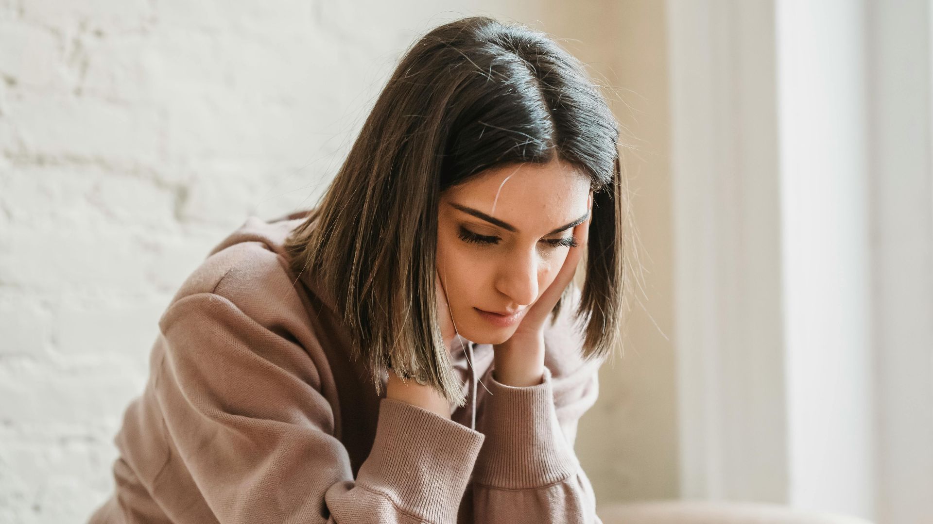 Unhappy female in casual wear touching face and looking down while sitting on sofa in light living room at home