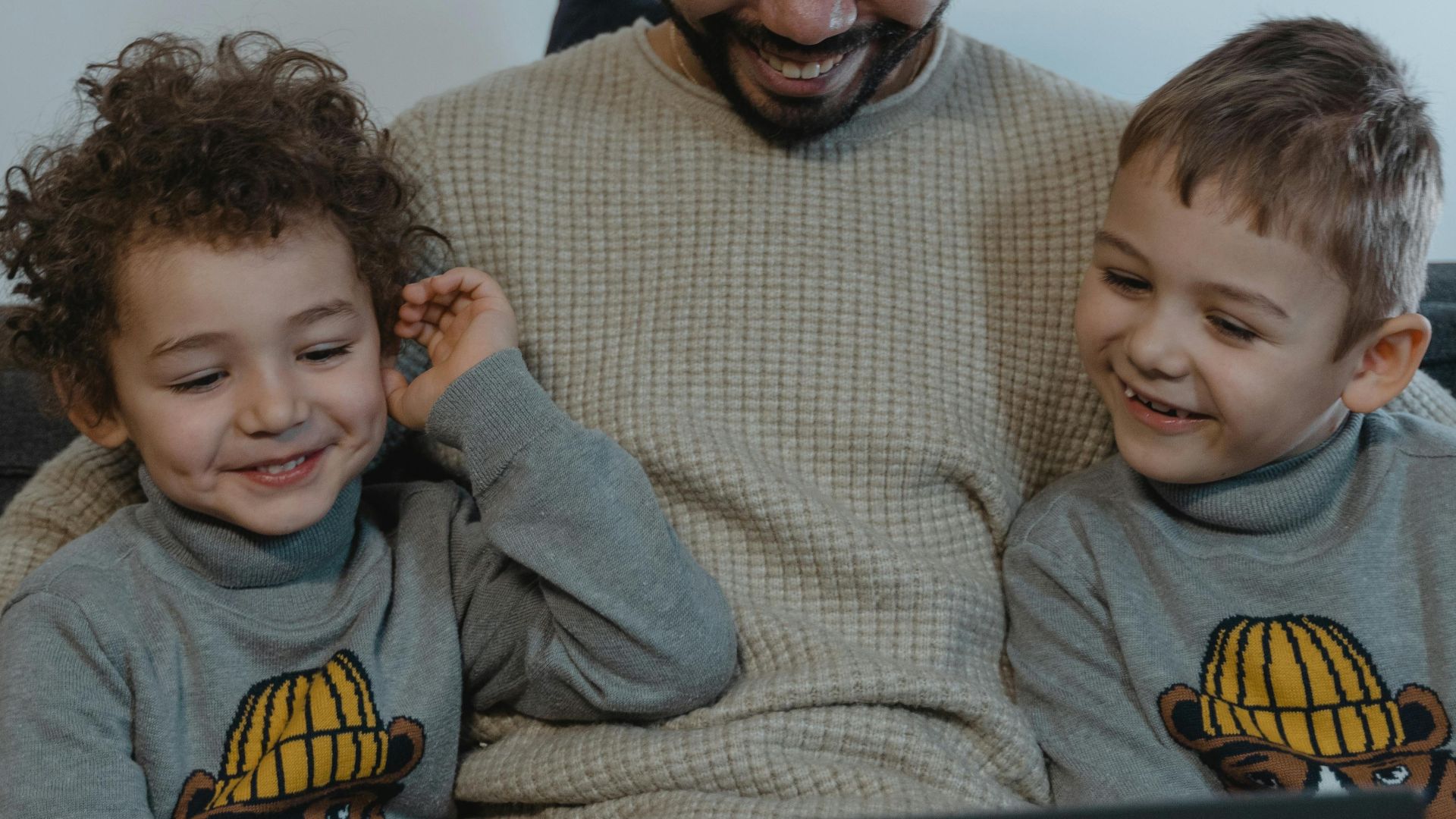 Smiling father and sons enjoying time together with a laptop indoors, wearing matching sweaters.