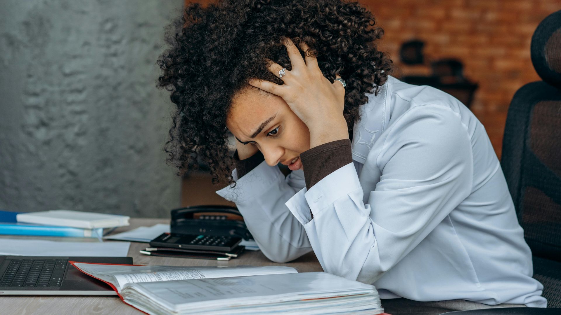 Woman sitting at desk appearing stressed while reviewing documents in an office setting.