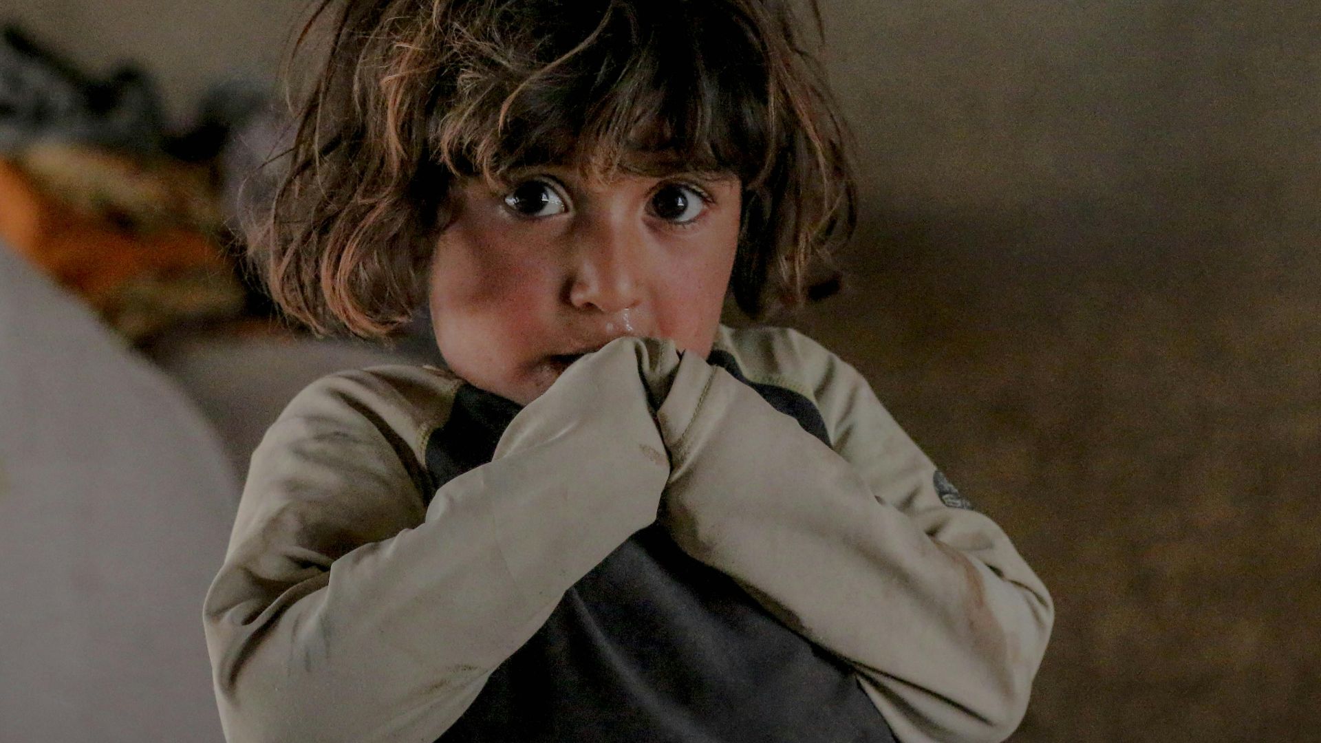 Little refugee child with dirty hair covering mouth with hands and looking at camera while standing in shabby room inside