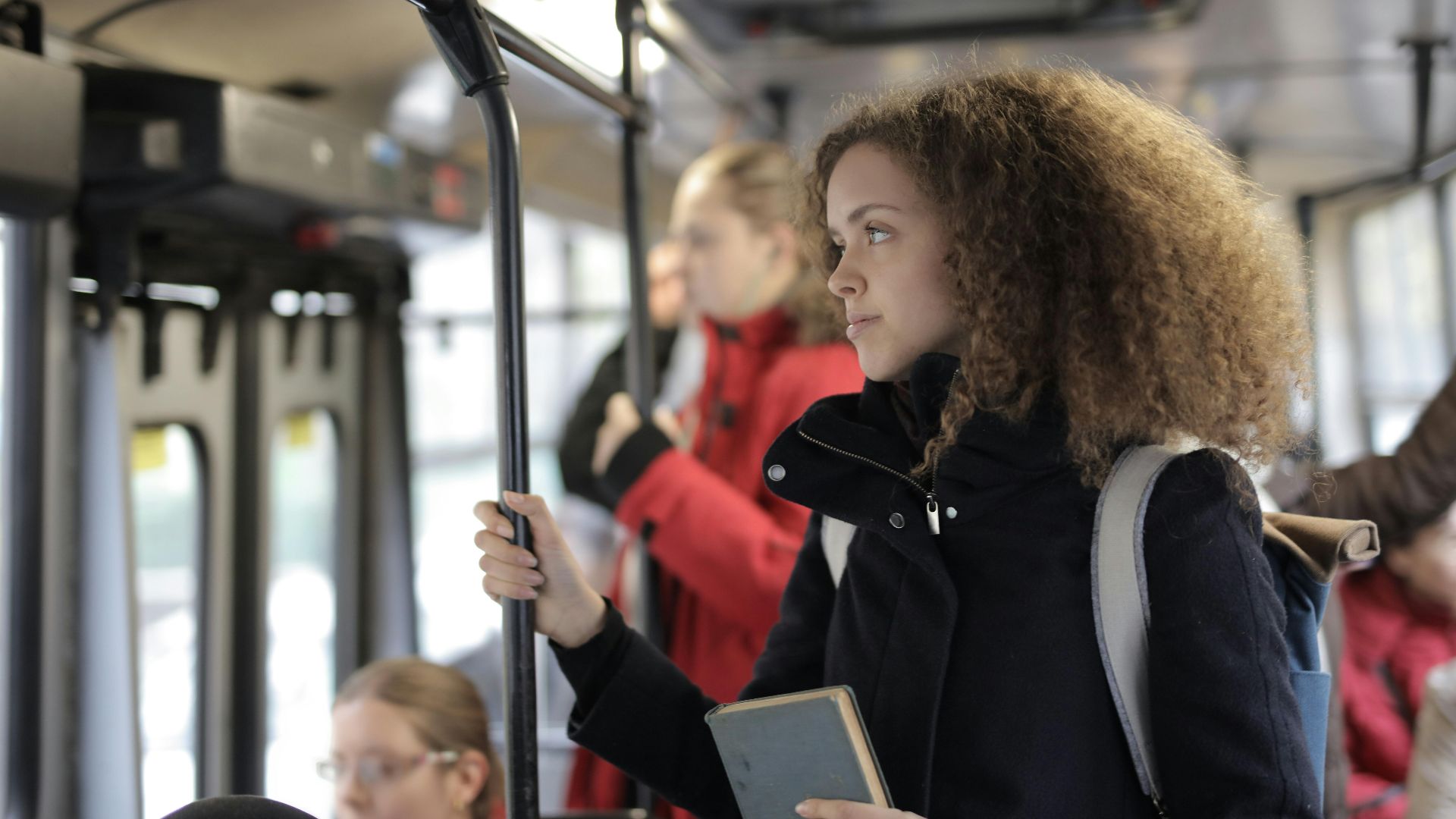 Side view of serious young lady with curly hair in casual warm coat with backpack and book riding bus during trip