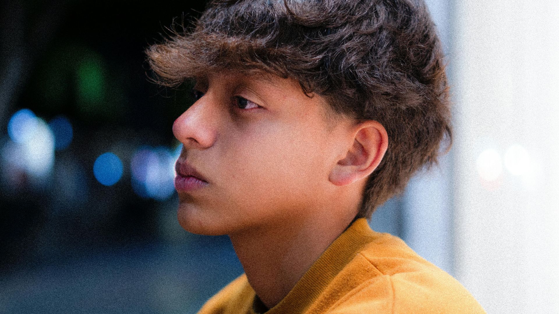 Profile portrait of a teenage boy in a contemplative mood, captured at night with vibrant urban bokeh.