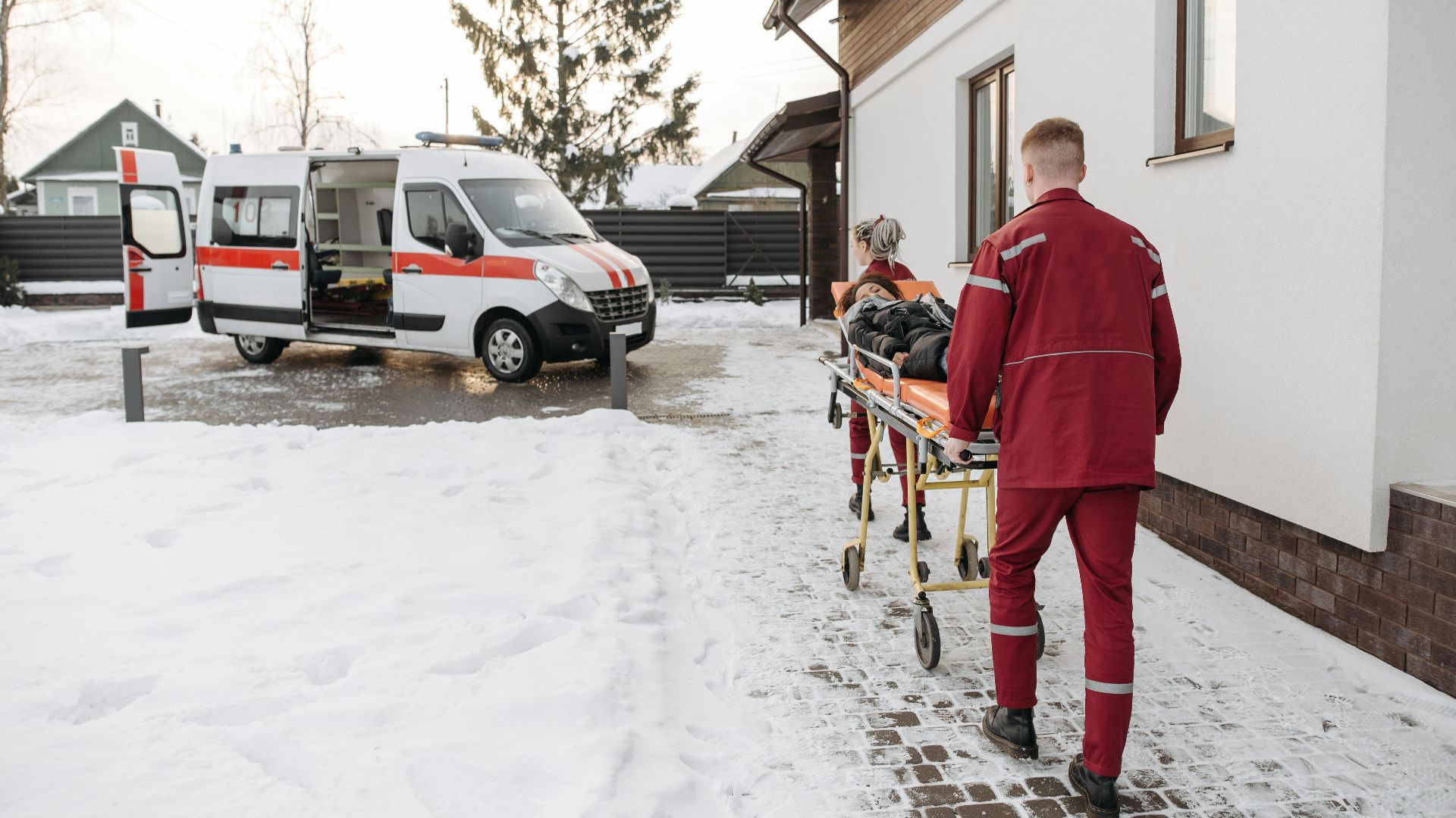 Paramedics transport a patient on a stretcher to an ambulance in a snowy residential area.