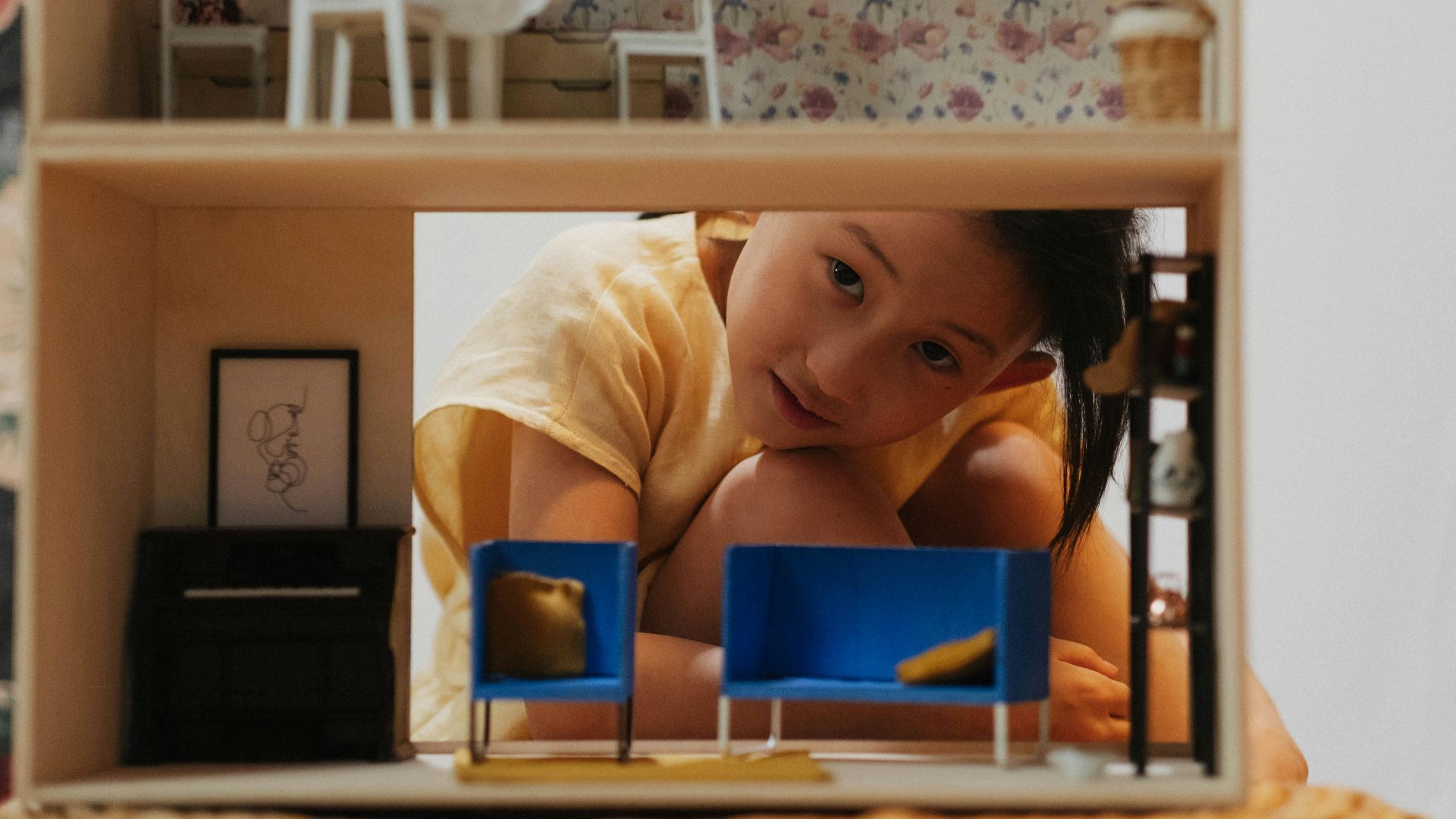 A young girl peers into a dollhouse, showcasing intricate miniature furniture in an indoor setting.