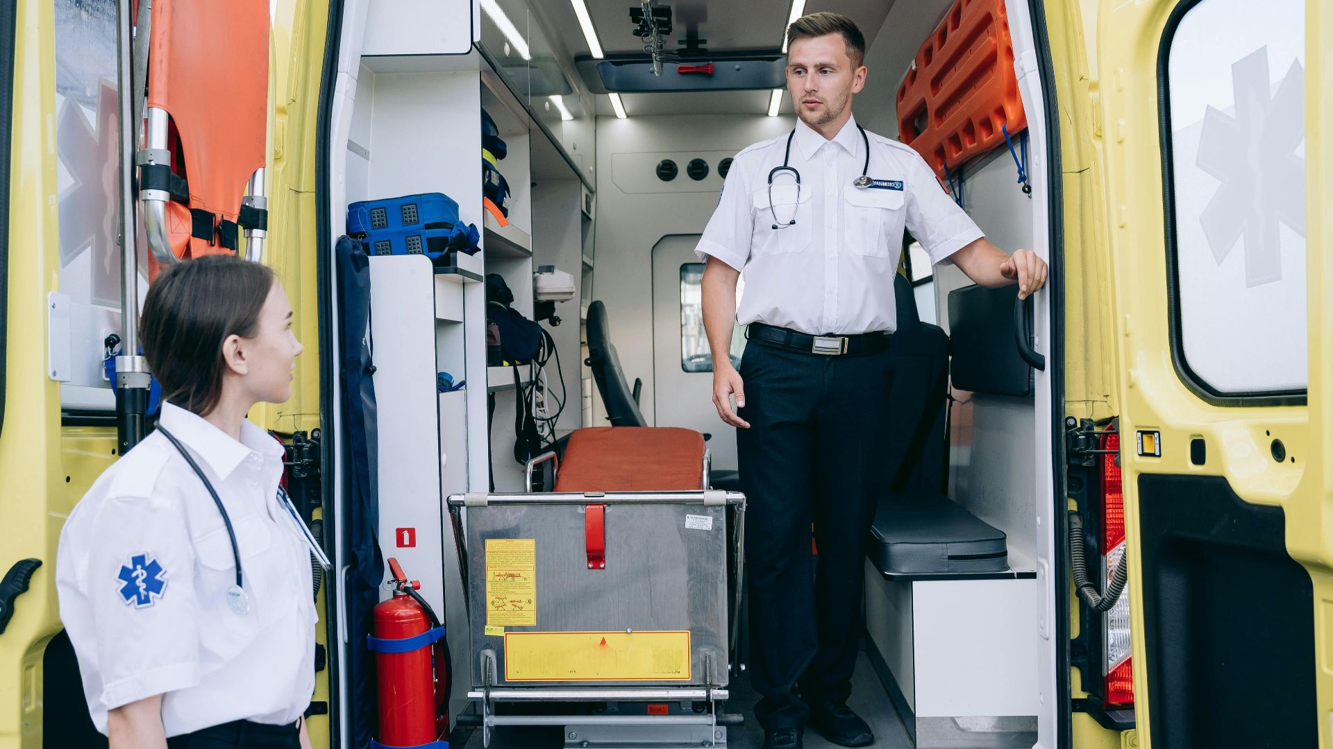 Two paramedics in uniforms and stethoscopes preparing ambulance for upcoming medical emergencies.