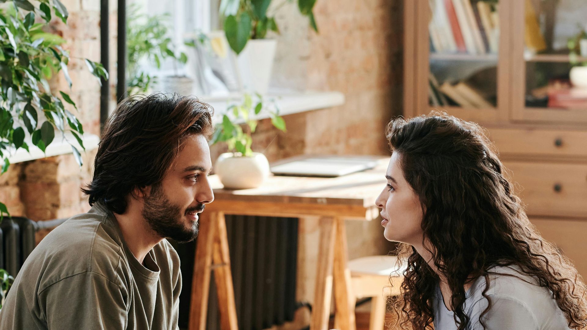 Romantic couple holding hands and talking in a cozy, plant-filled bedroom.