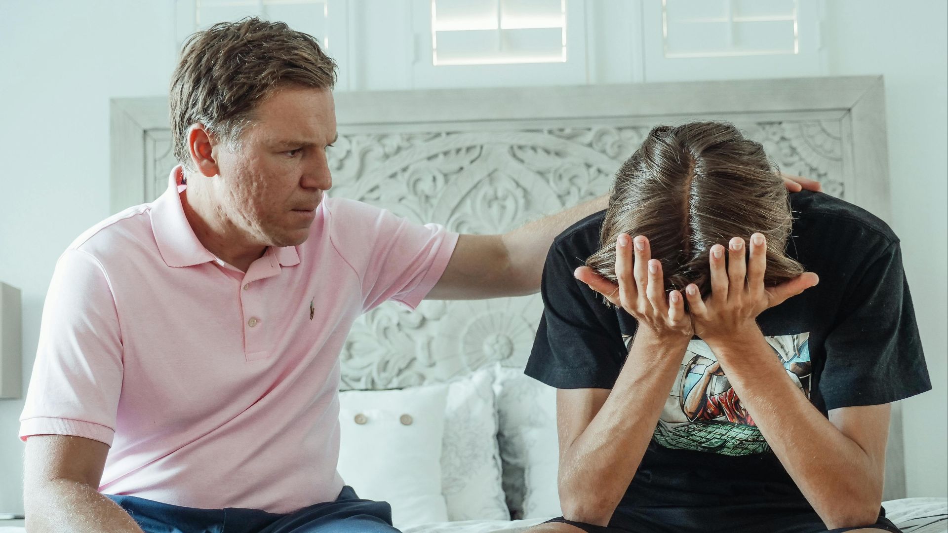 A compassionate father consoles his upset teenage son on a bed indoors.
