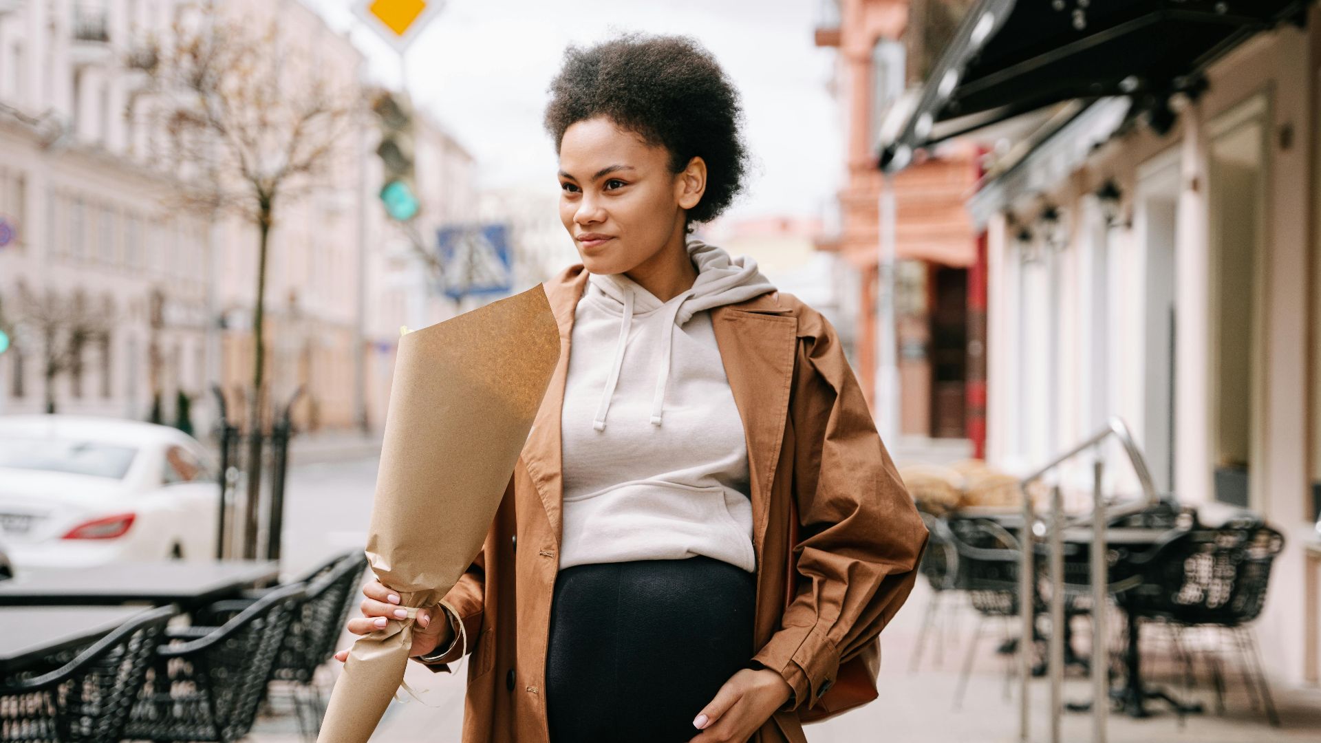 A stylish pregnant woman with afro hair holding flowers, walking down a charming city street.