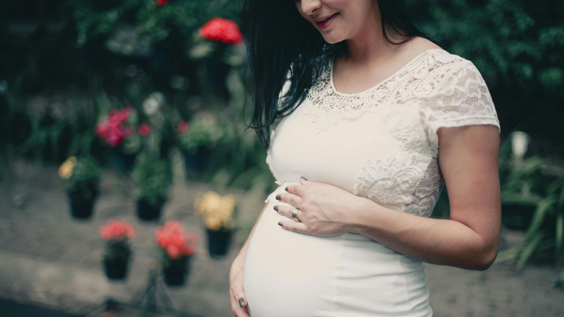 Pregnant woman in white lace dress, posing outdoors with flowers, exuding happiness.