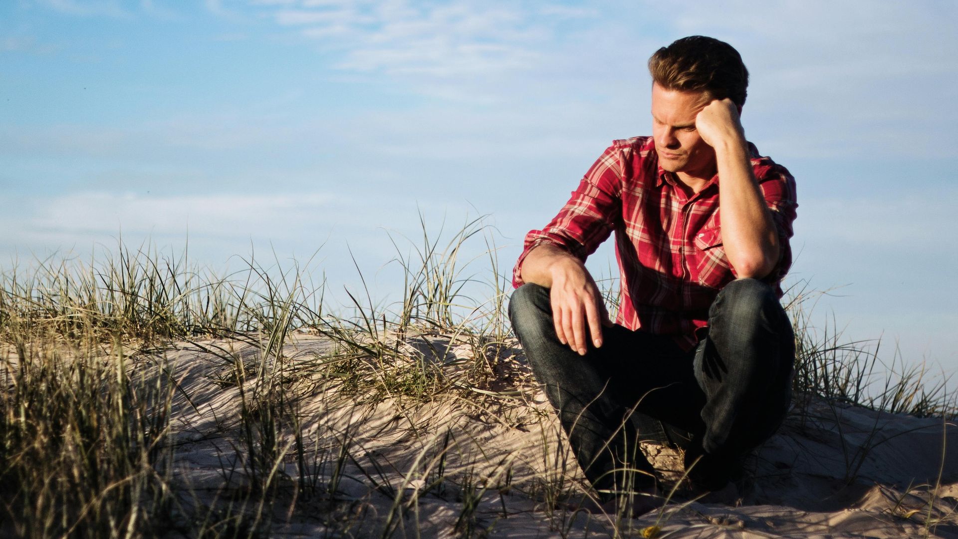 Man in a red plaid shirt sitting thoughtfully on a sandy dune in Australia, surrounded by grass.