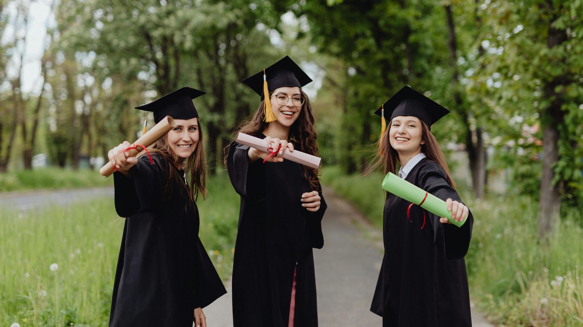 Young women in graduation gowns celebrating with diplomas in a park.