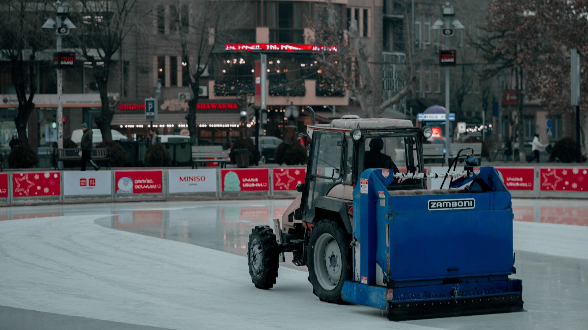 A Zamboni machine resurfacing an outdoor ice rink in a city during winter.