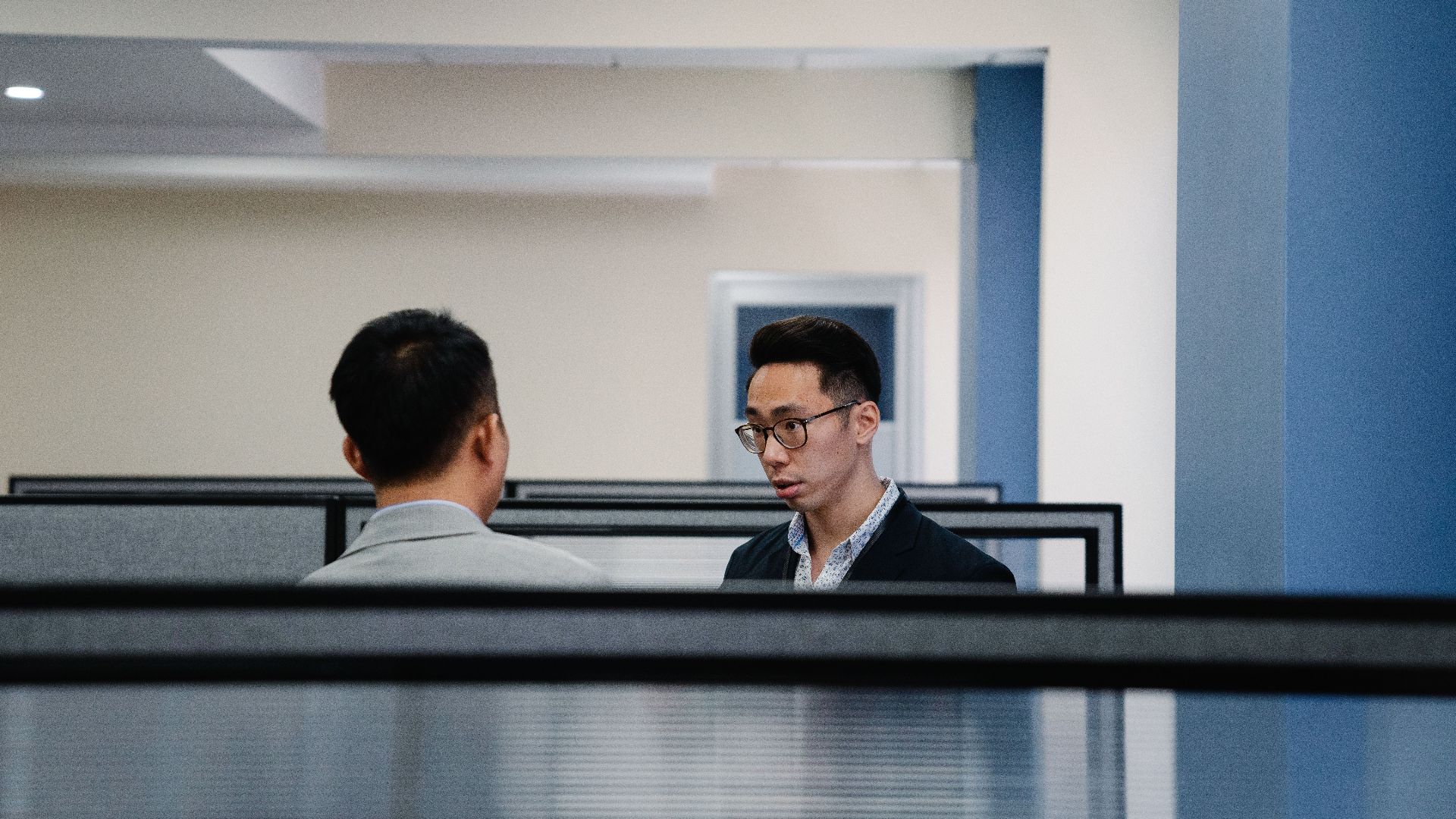 Two professionals engaged in conversation within an office cubicle setting.