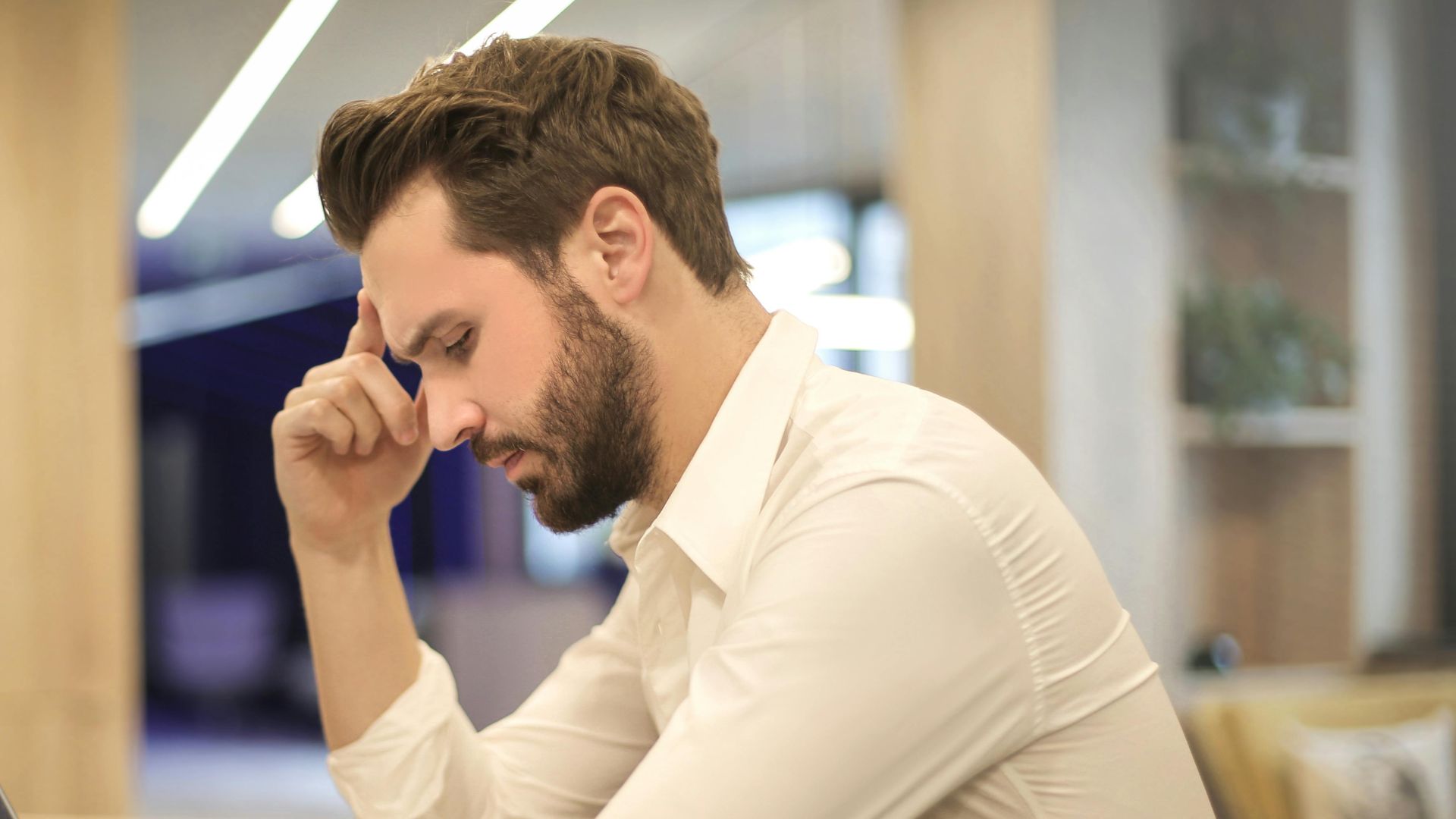 A thoughtful man in a shirt works on his laptop at a modern indoor office space.