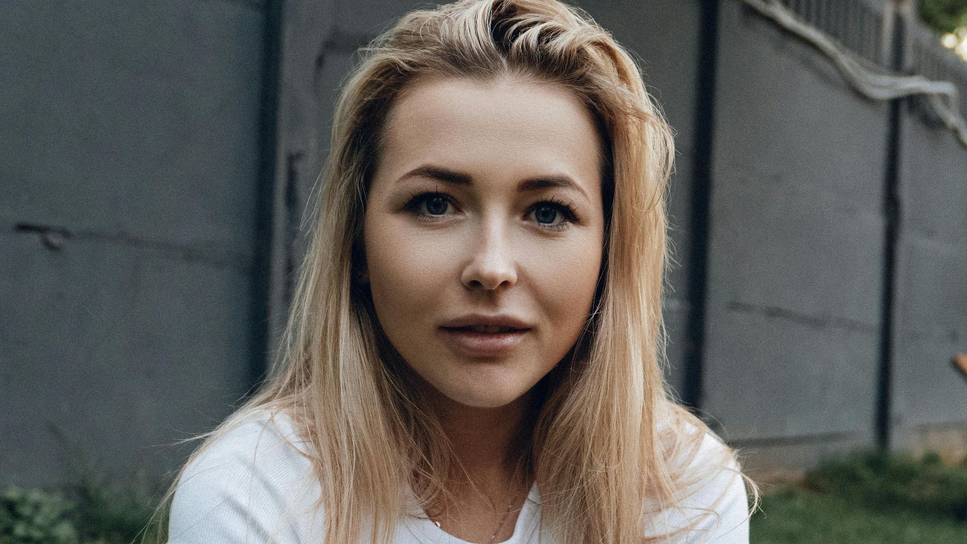 Young woman with blonde hair sitting outdoors, wearing a casual white shirt.