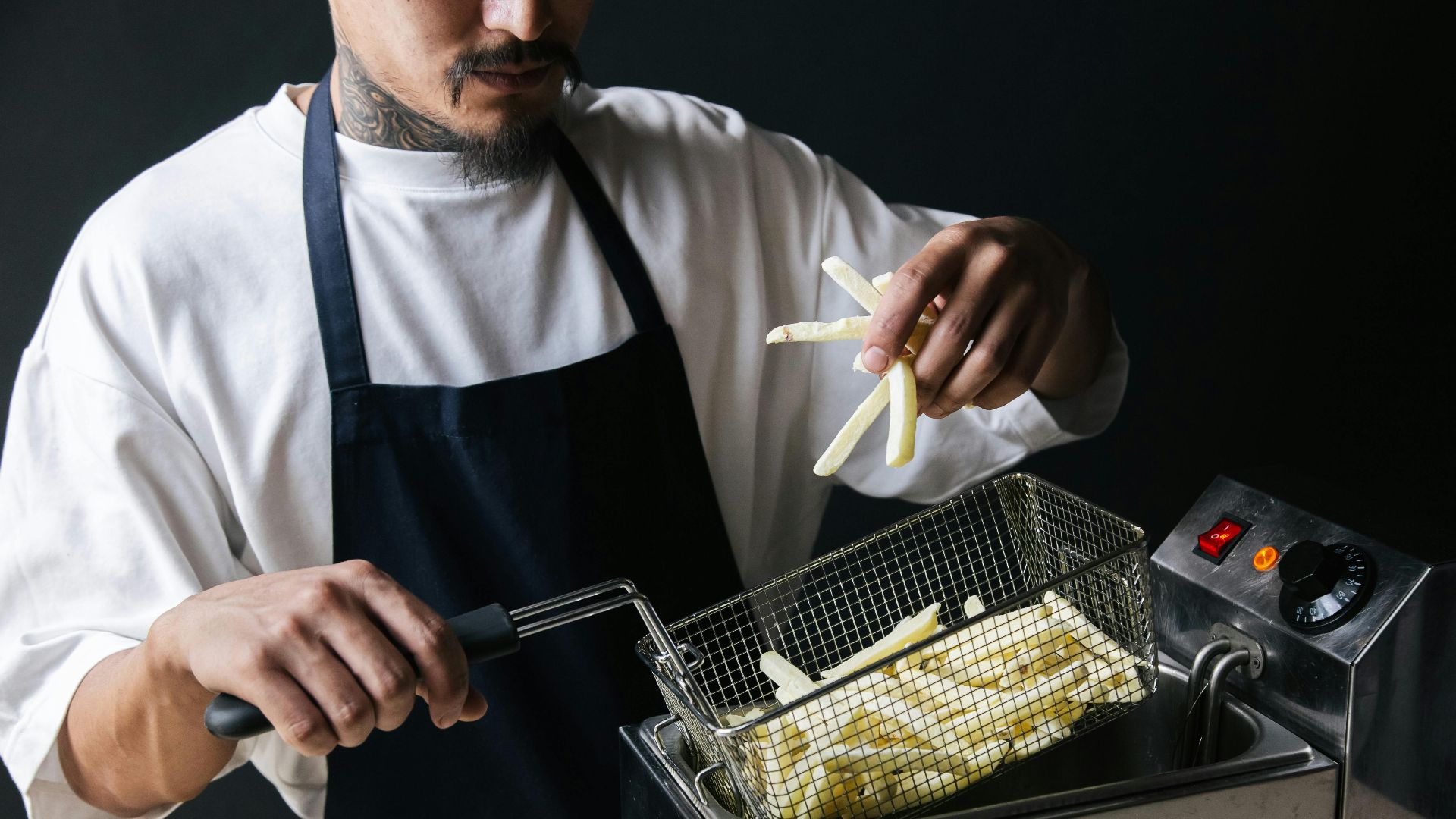 A chef handling raw french fries with a strainer basket in a deep fryer.