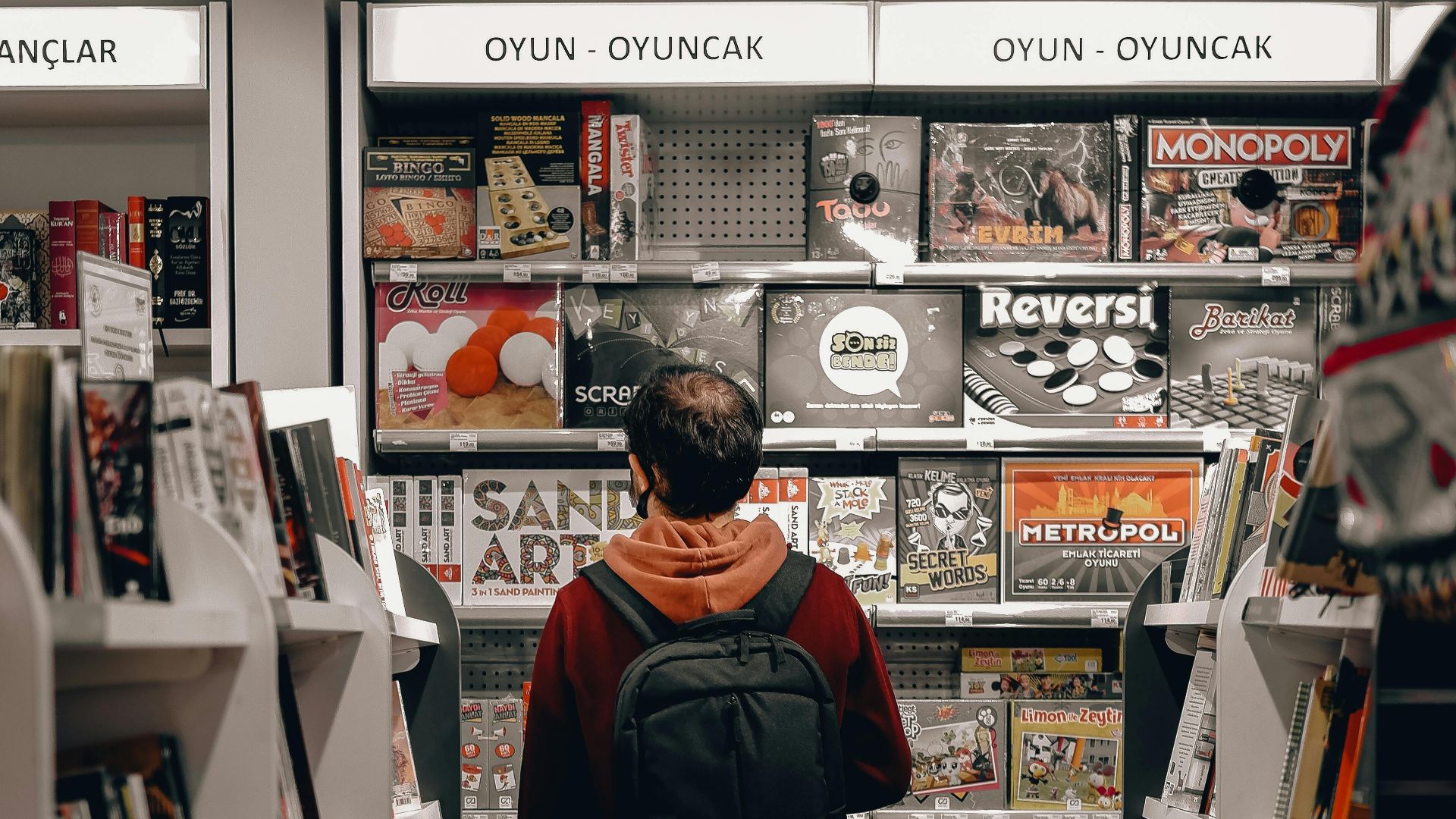 Man with a backpack browsing board games in a store aisle filled with books and games.