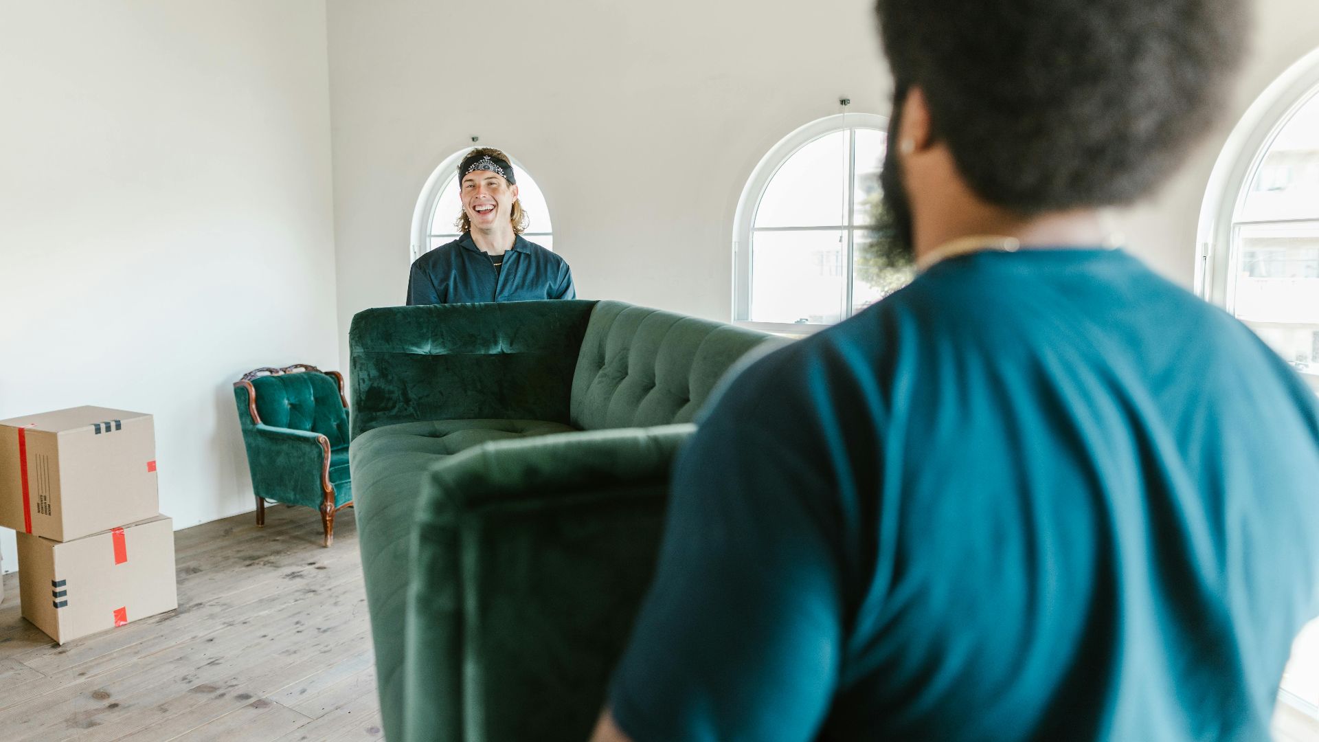 Two movers carrying a green couch in a sunlit room with packed boxes.