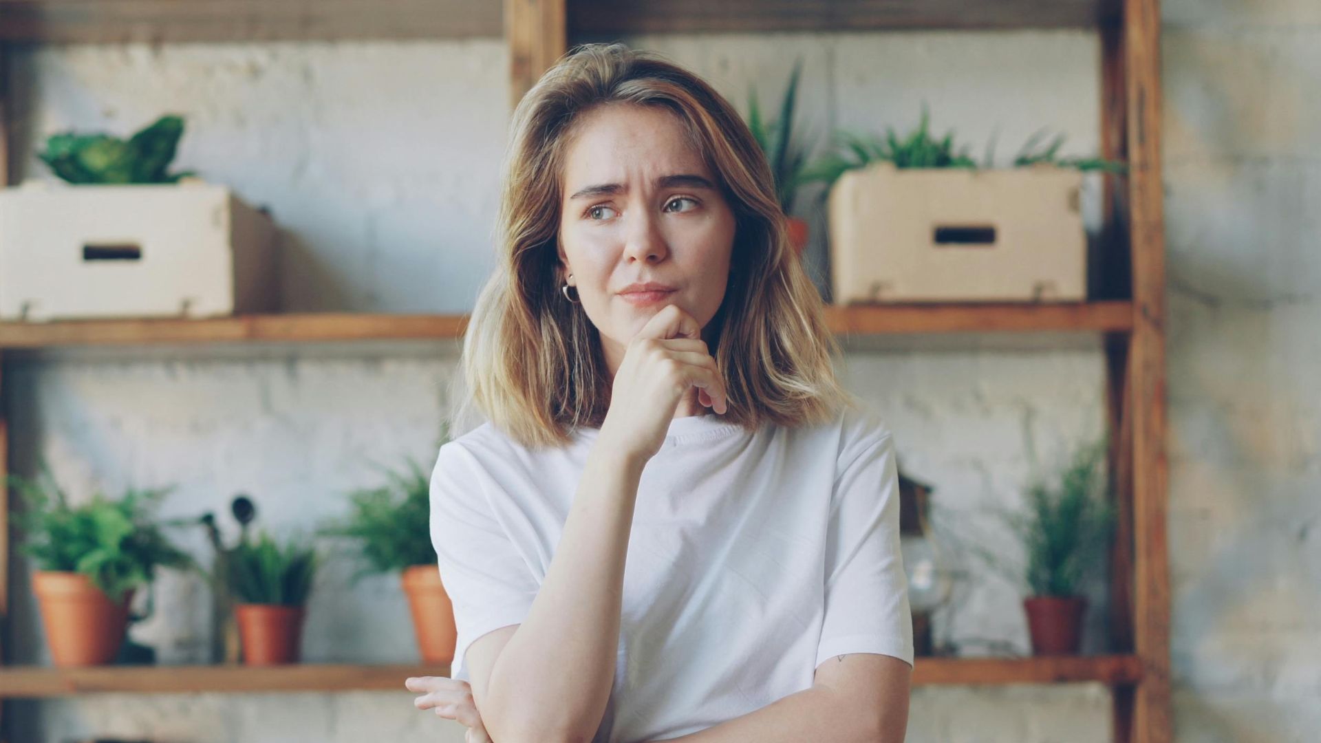 Pensive young woman in casual attire stands in a stylish room with plants.