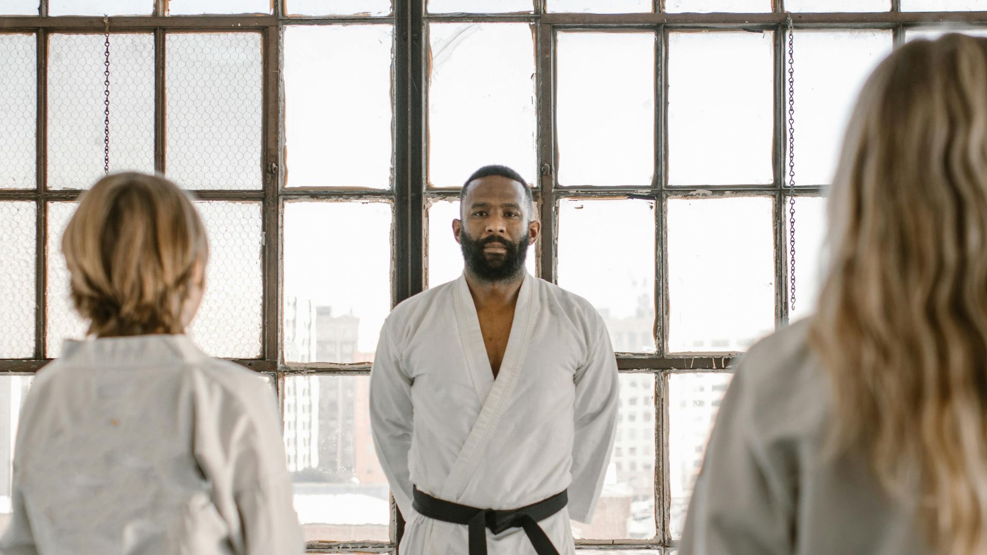 Martial arts instructor and students practicing in a well-lit dojo with large windows.