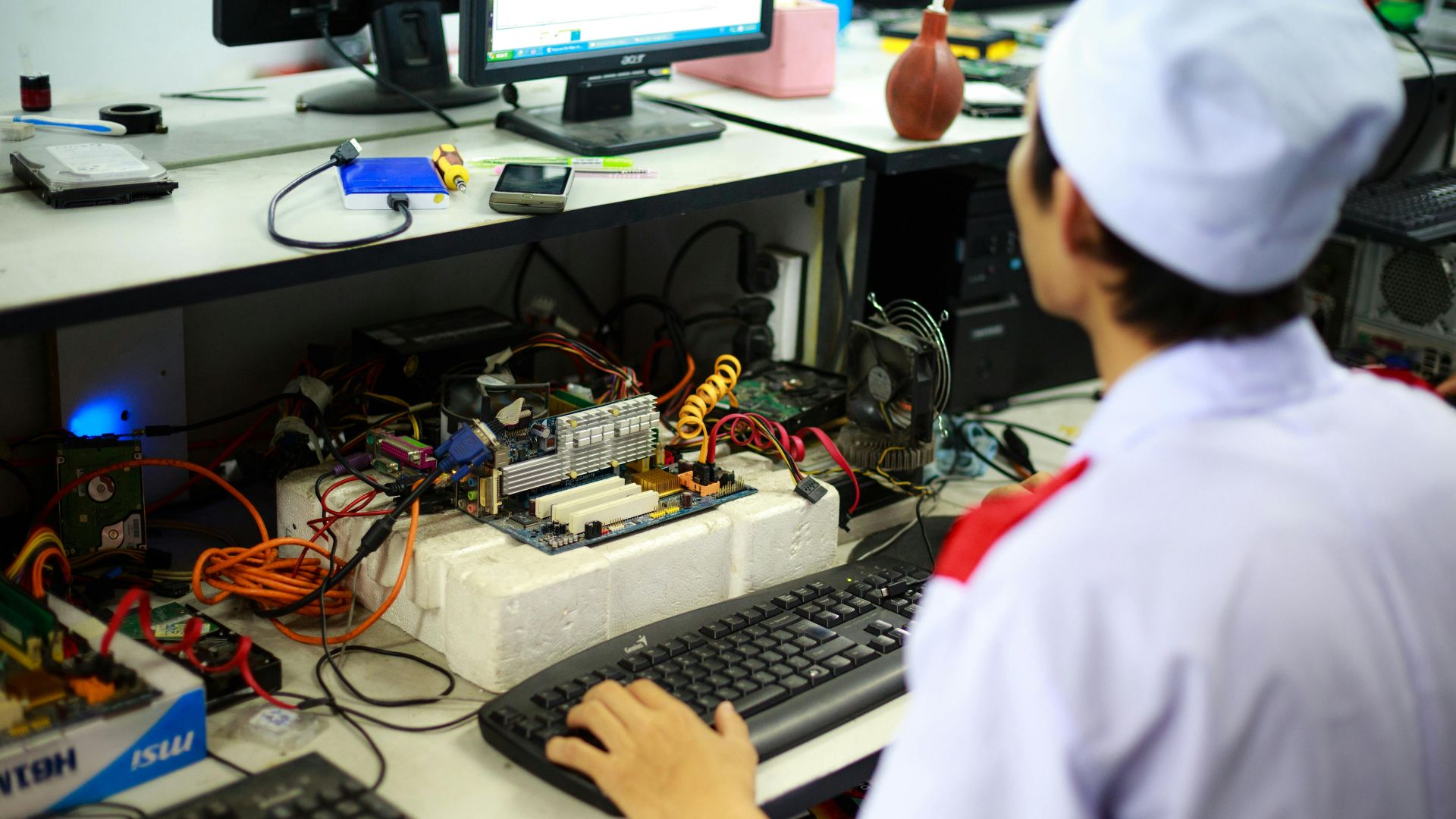A technician in uniform working on a motherboard at a desk in a laboratory setting.