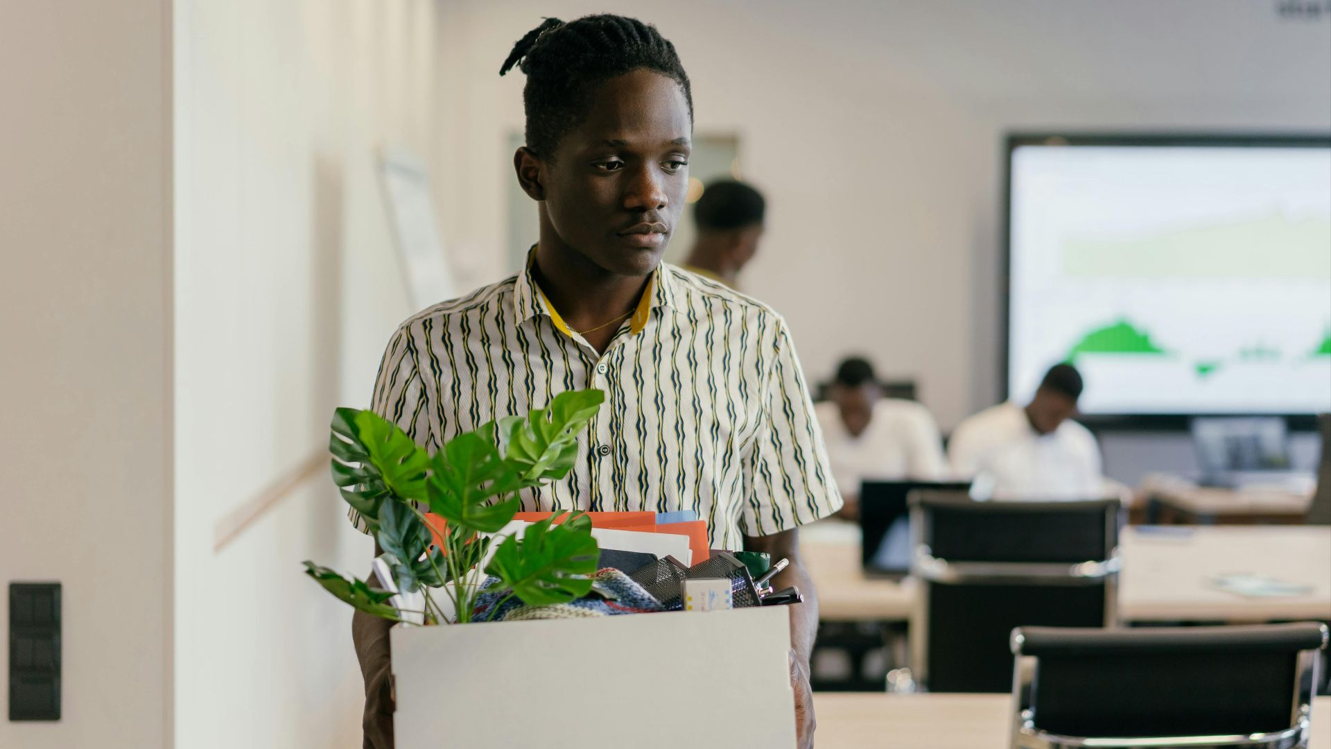 A serious young man carrying a box of personal items in an office setting, suggesting dismissal or resignation.