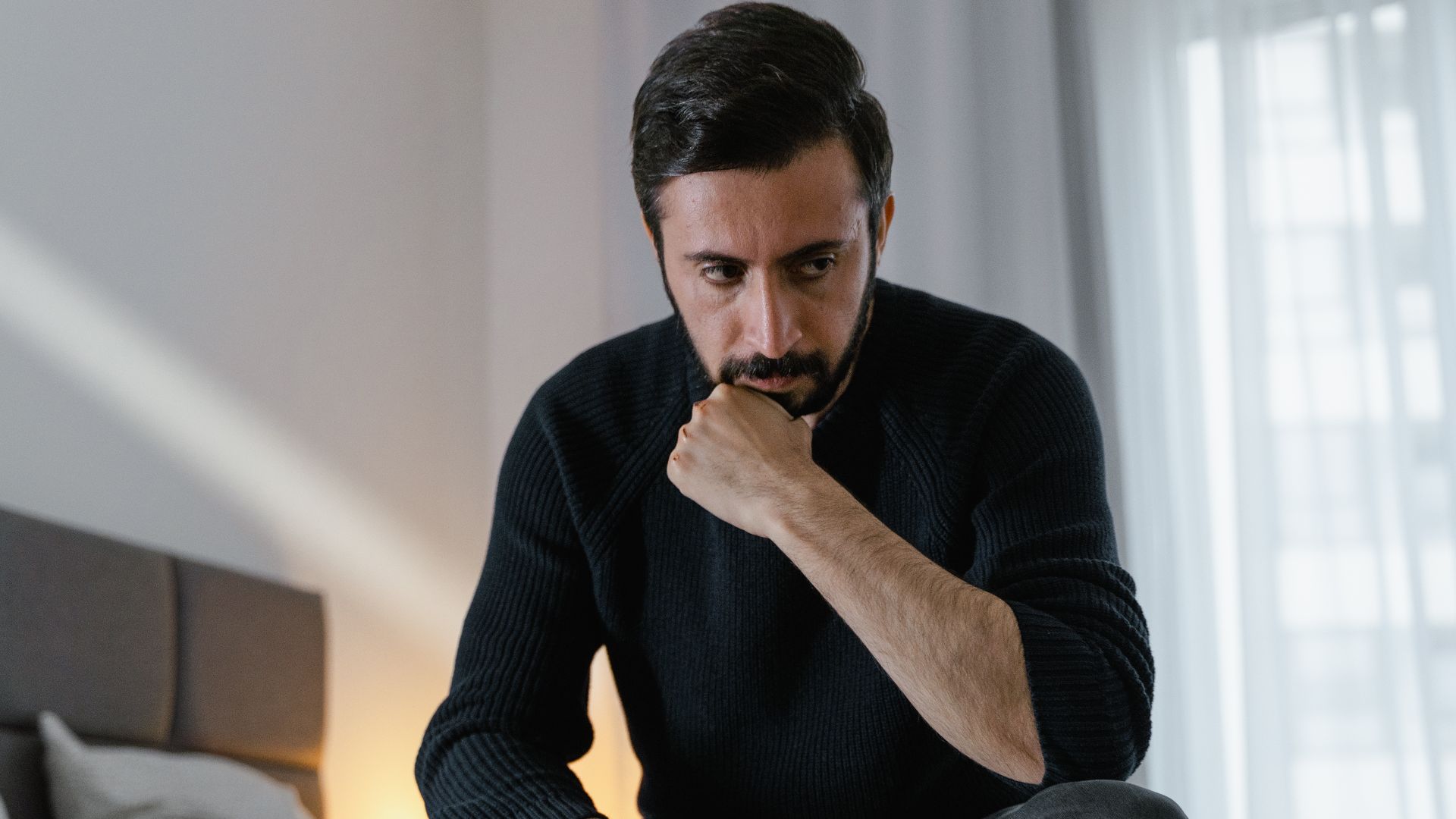 A thoughtful man sits on a bed in a modern bedroom, hand resting under chin.